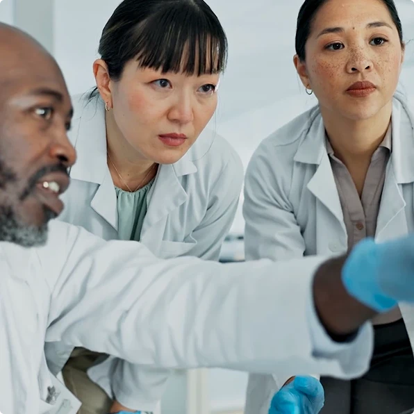 Three scientists in lab coats examining data on a screen or sample closely. The group includes a man and two women, all focused intently, in a modern laboratory setting.
