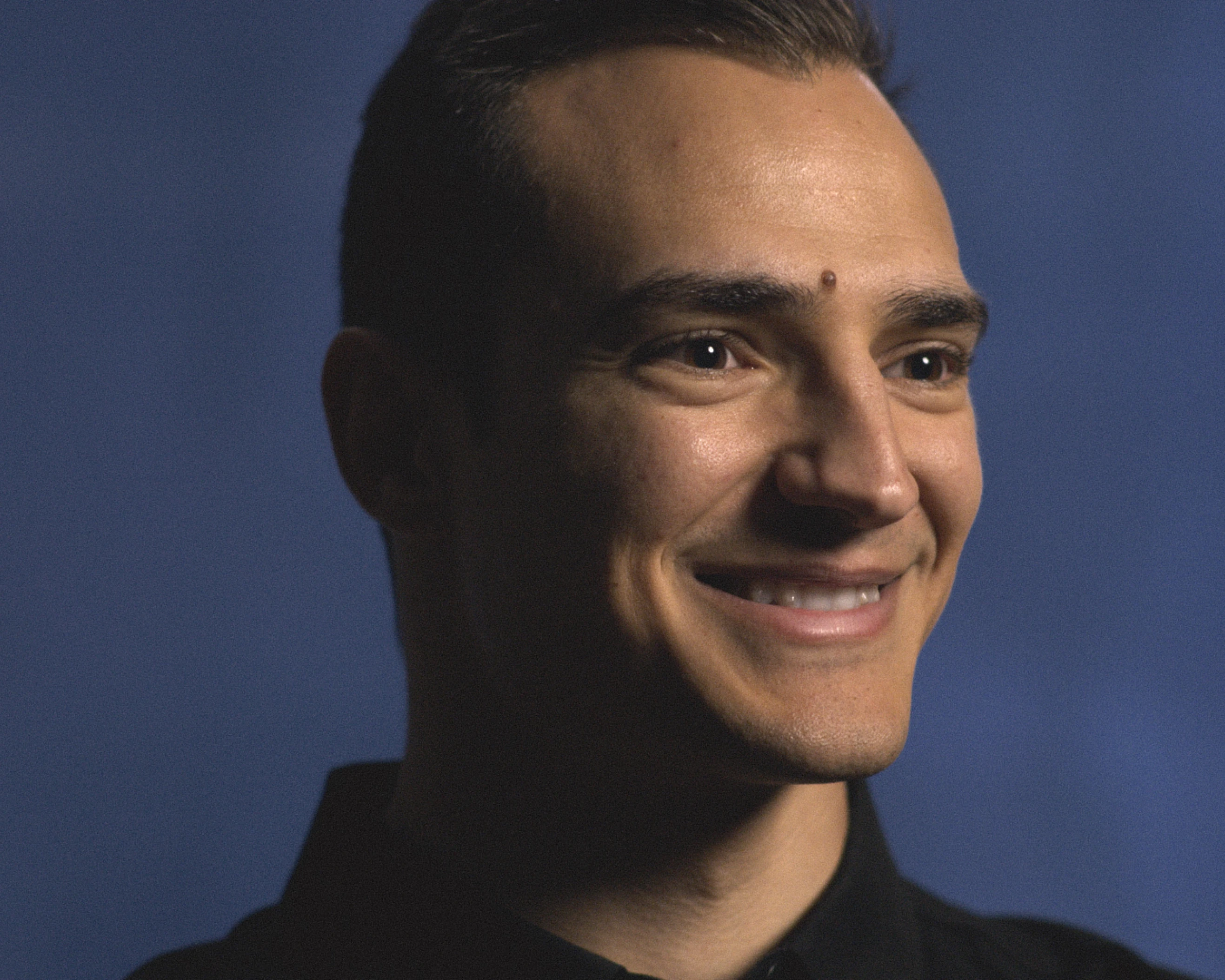 Close-up portrait of a young man smiling against a plain blue background, wearing a dark collared shirt.