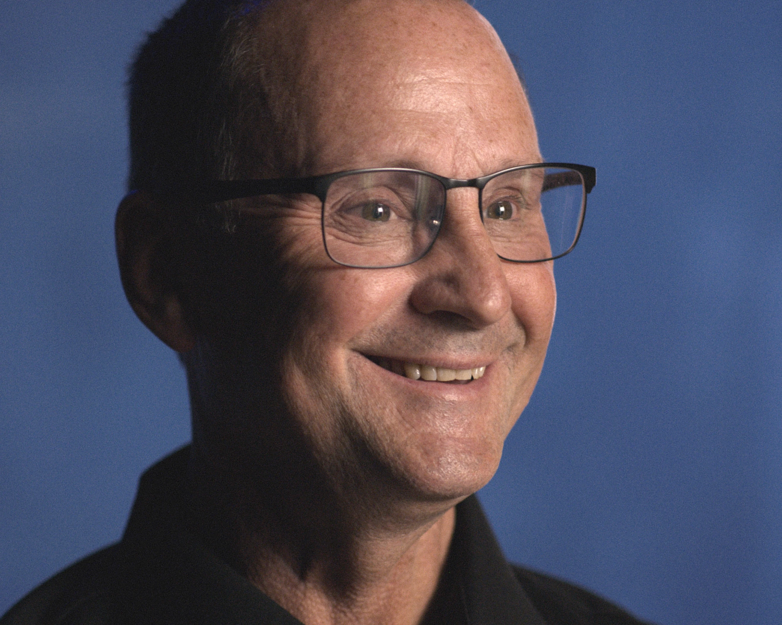 Close-up of an older man with a bald head wearing glasses and a dark shirt, smiling against a plain blue background.
