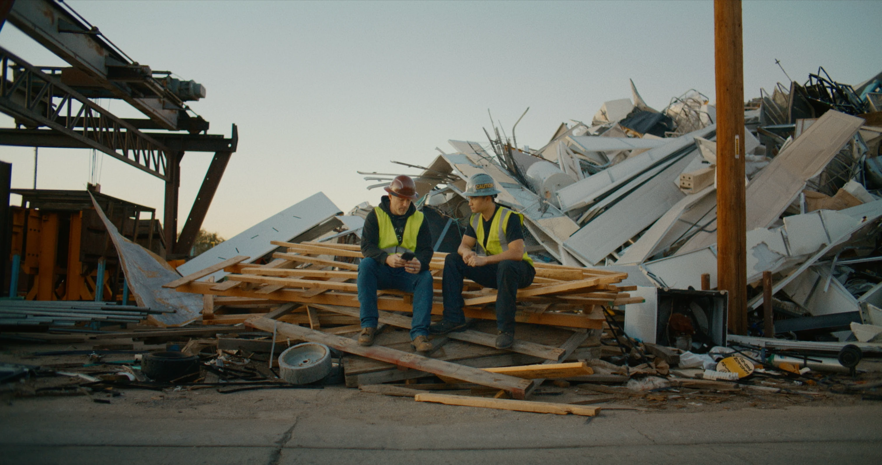 Two construction workers wearing hard hats and safety vests sit on stacked wooden pallets, talking in front of a large pile of scrap metal at an industrial yard.