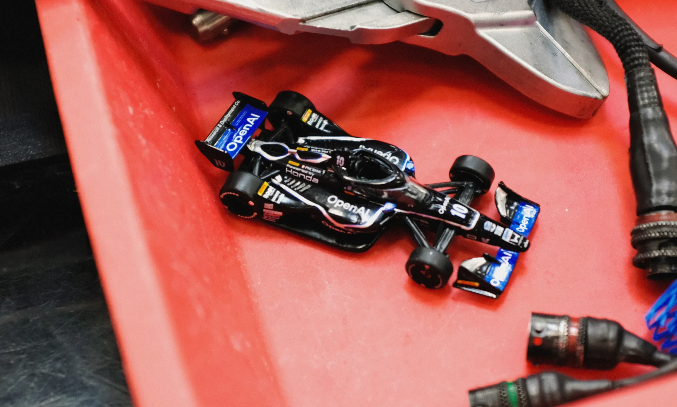 An OpenAI-branded model IndyCar on a red tray beside workshop tools.