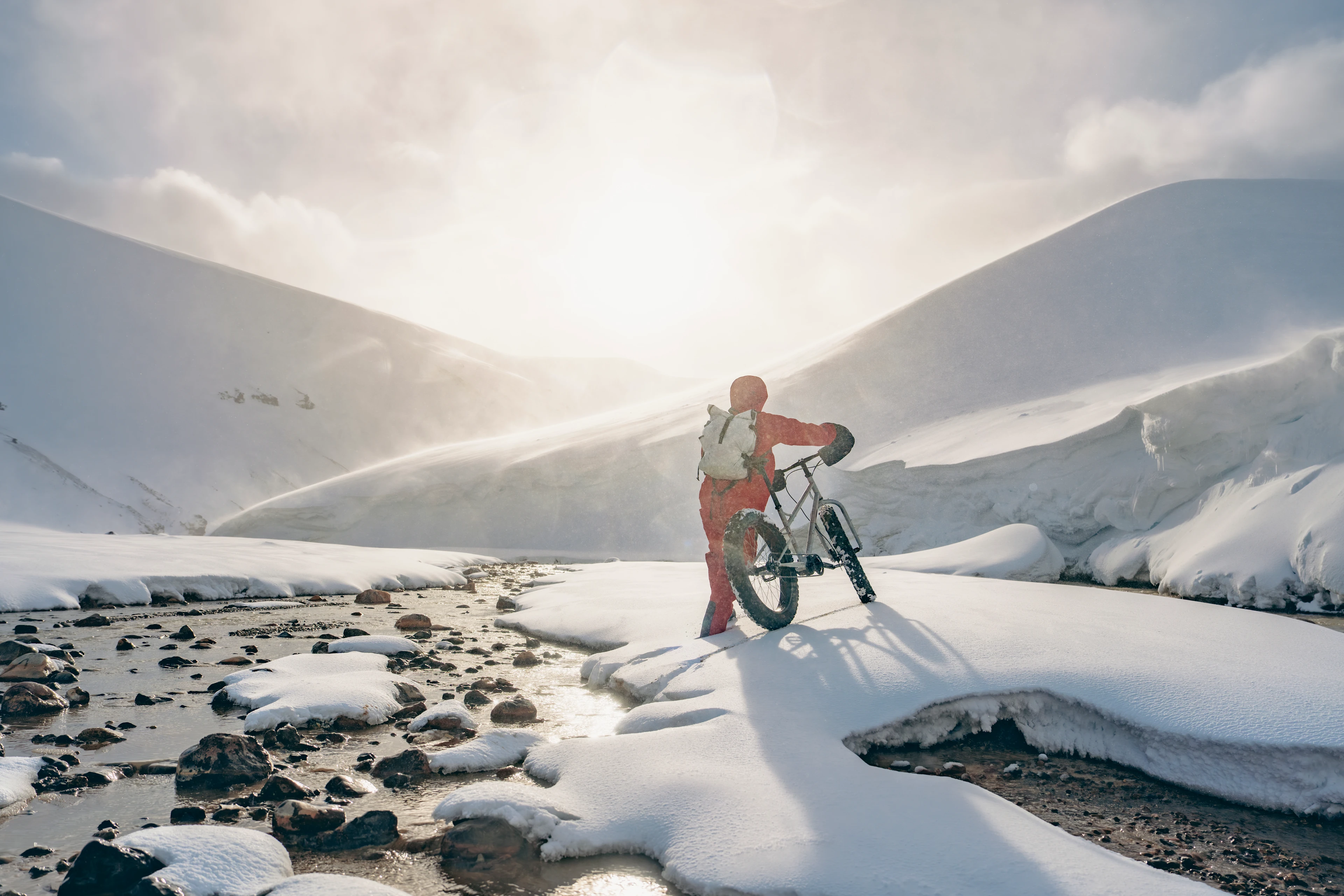 A cyclist stands with a fat-tire bike in a bright snowy mountain landscape.
