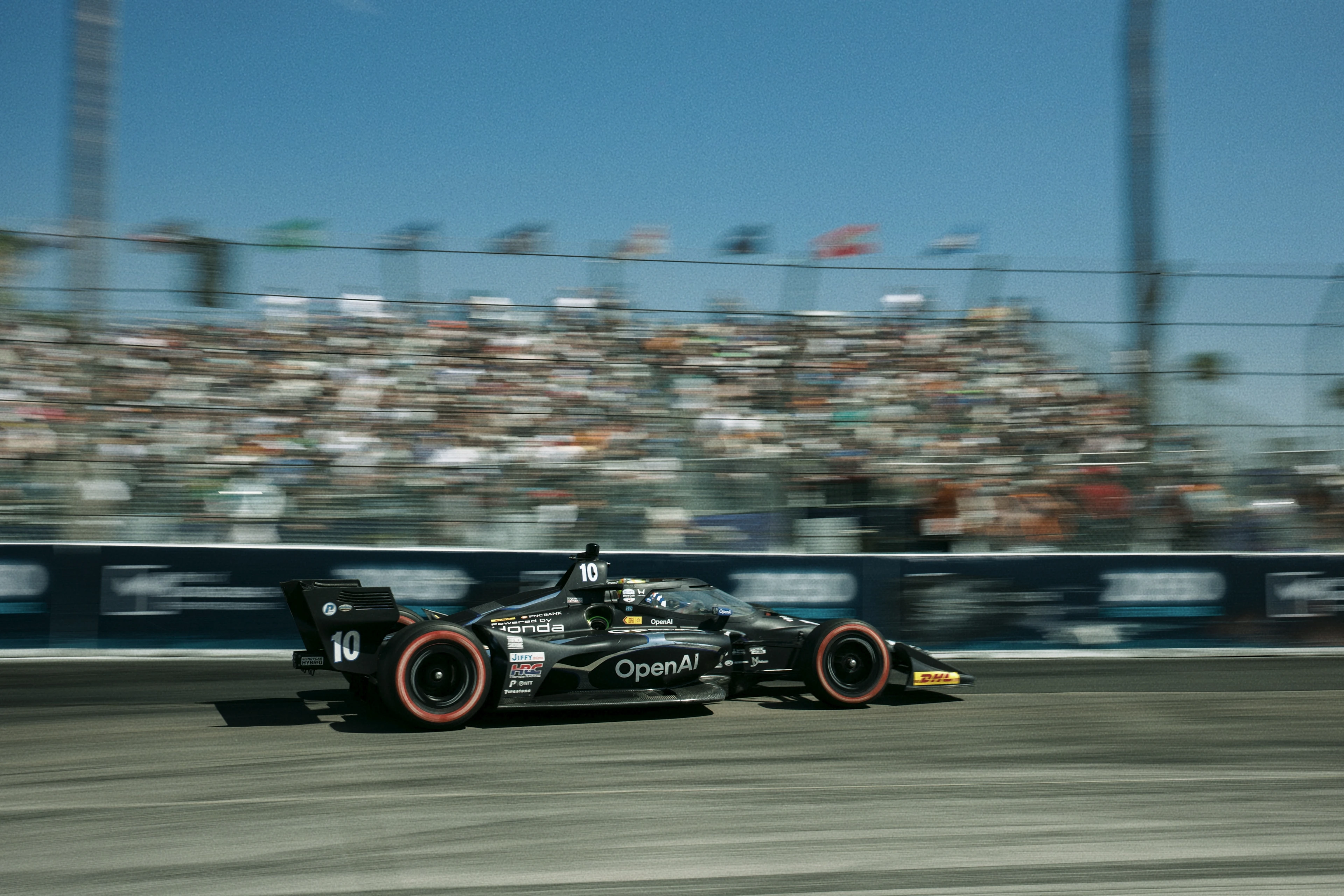 A black Formula 1 race car with the number 10 and OpenAI logo speeds past a grandstand filled with blurred spectators under a clear blue sky.