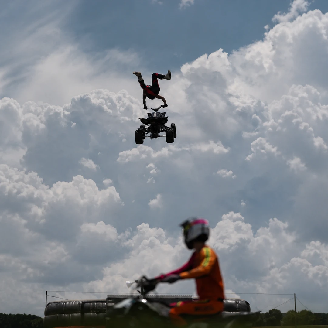 An ATV Big Air Tour rider performs an airborne stunt against a cloudy sky.