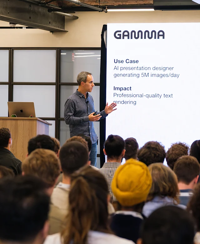 A presenter stands on stage giving a talk to a seated audience. They are gesturing with one hand while holding a clicker in the other, with a laptop on a podium nearby. A large screen next to them displays the word “GAMMA” with text beneath it reading “Use Case: AI presentation designer generating 5M images/day” and “Impact: Professional-quality text rendering.” The audience is visible in the foreground, facing the speaker. The setting has an industrial ceiling with exposed beams.