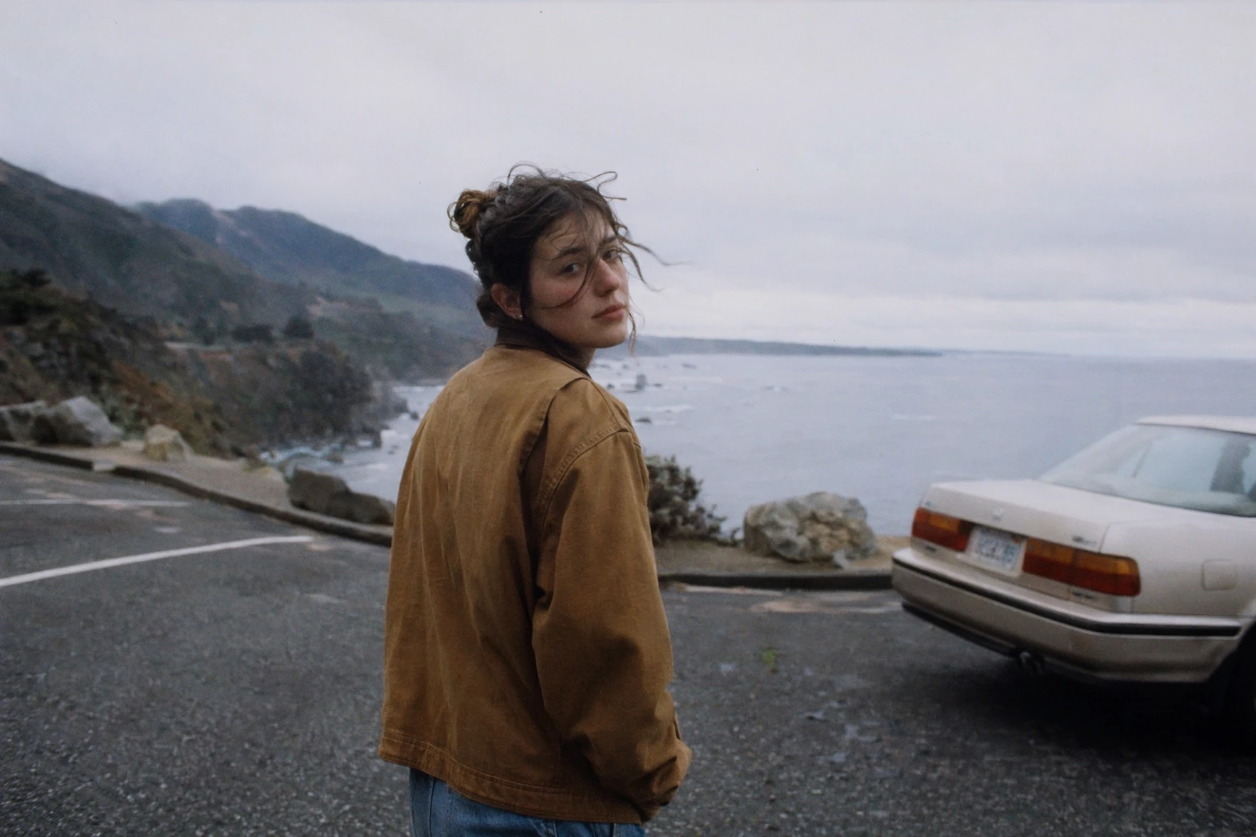 Generated with ChatGPT Images 2.0, this cinematic candid portrait shows a person in a brown jacket looking back toward the camera at a coastal roadside overlook, with misty cliffs, ocean water, and a parked car under an overcast sky.