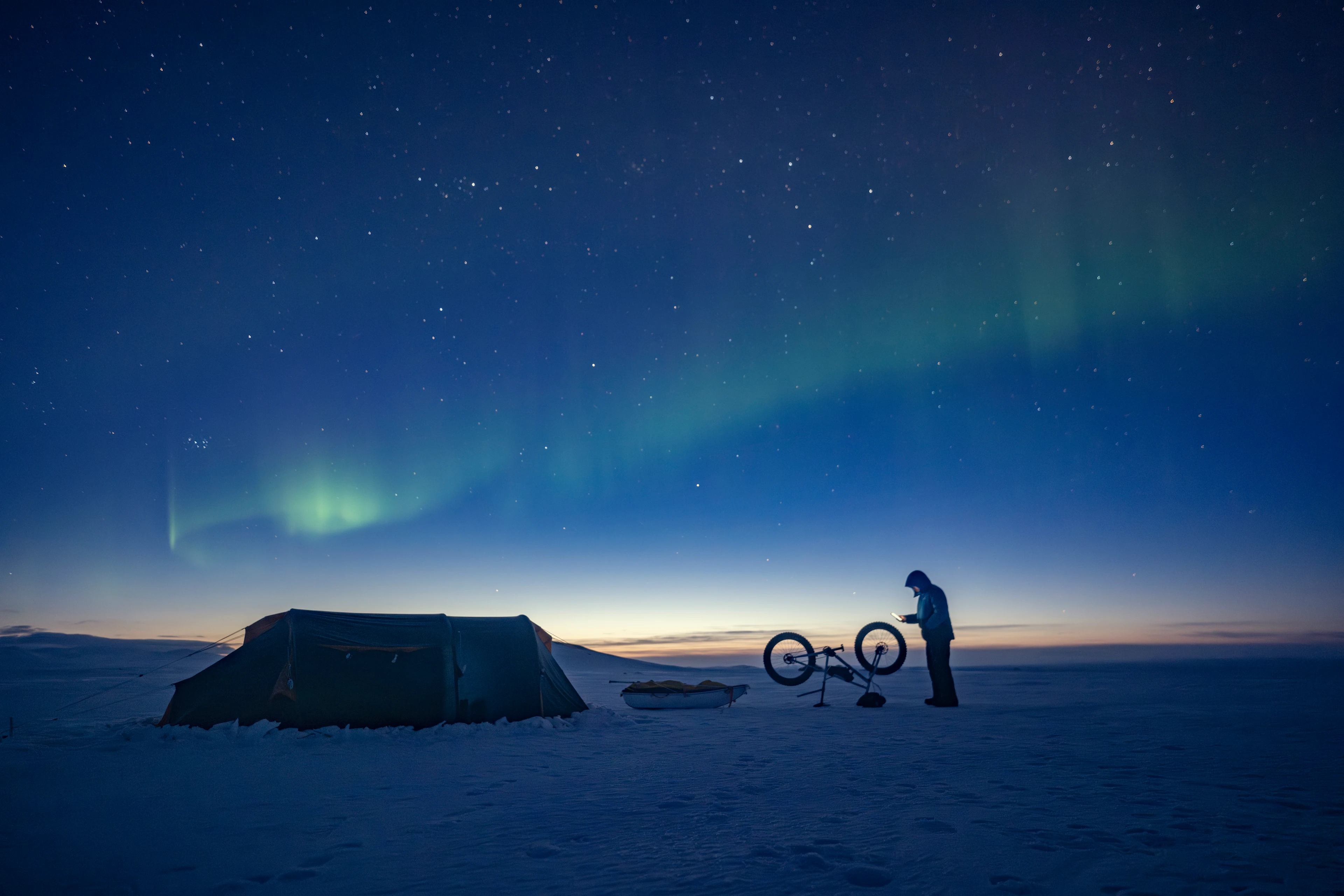 James stands beside his bike and tent under the northern lights during a remote training expedition in Iceland.