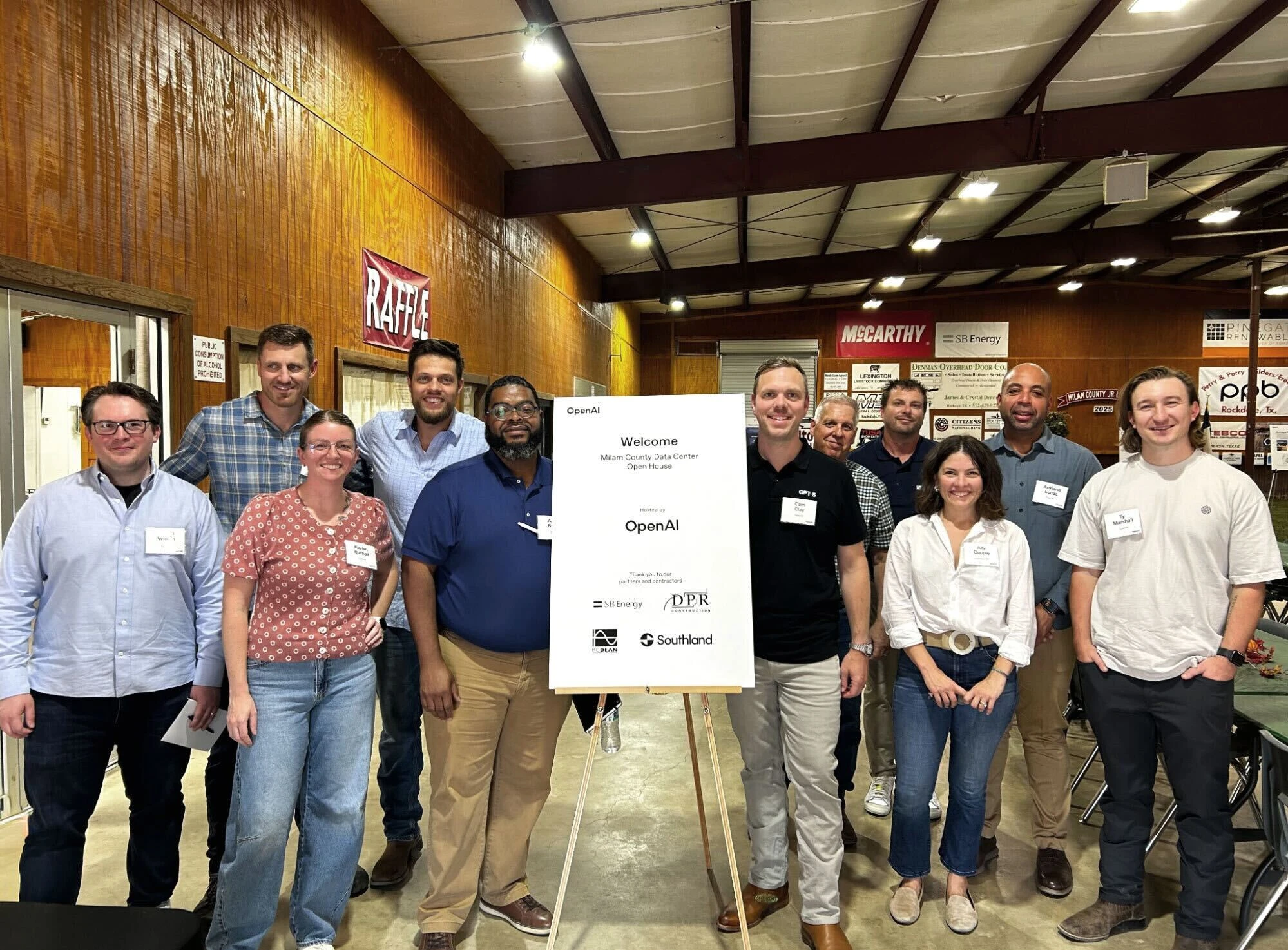 Group photo of Stargate community members from various partner companies standing together indoors around a welcome sign for the Milam County Data Center Open House hosted by OpenAI. Everyone is smiling in a casual, workshop-style event space.