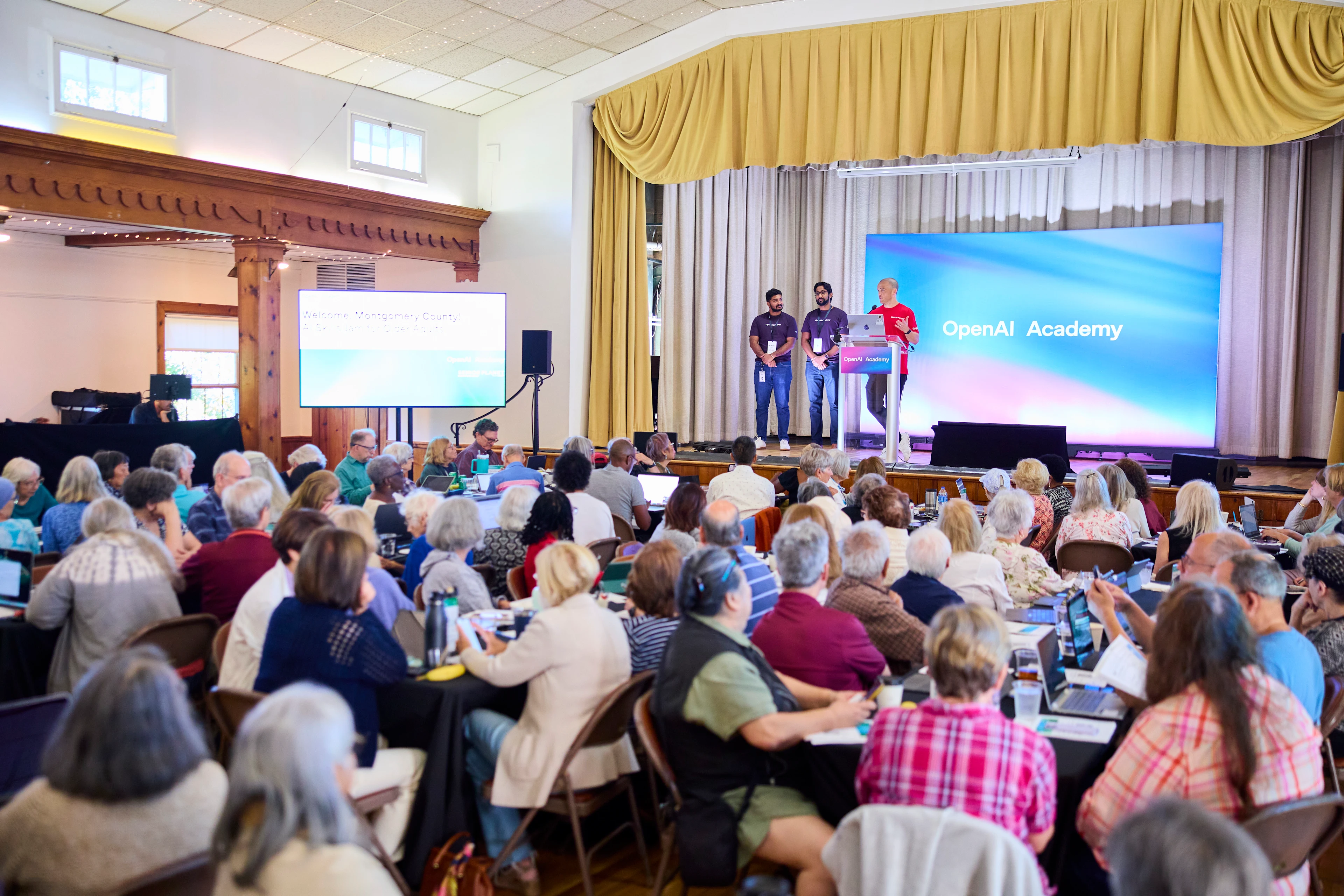 Presenters stand onstage before a large audience at an OpenAI Academy event.