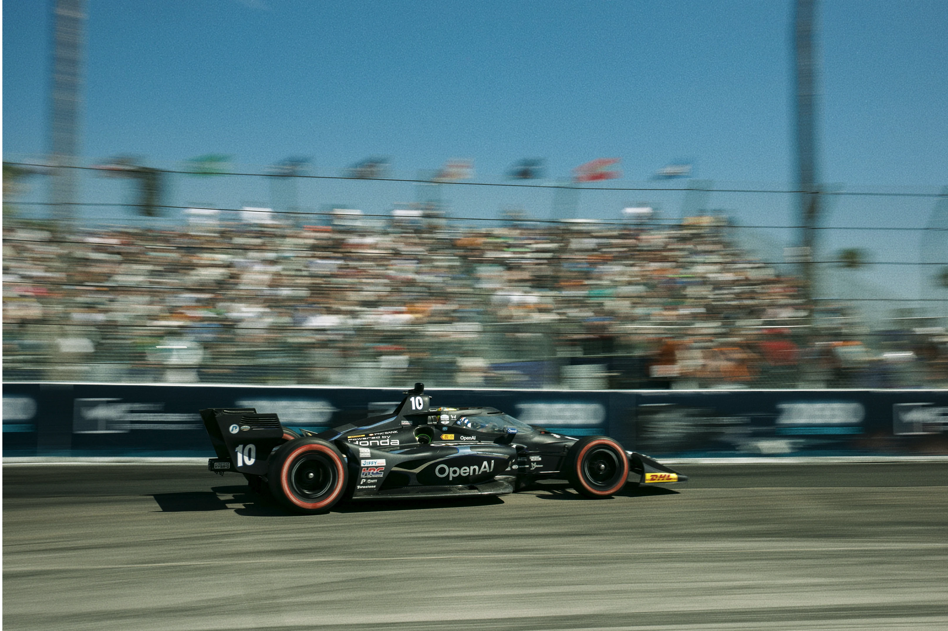 OpenAI-branded Chip Ganassi Racing car speeding past a grandstand.