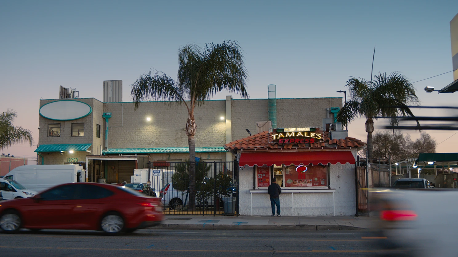 A small street-side tamale stand with a red awning and a neon ‘Tamales Express’ sign is lit at dusk, with palm trees behind it and blurred cars passing in the foreground.
