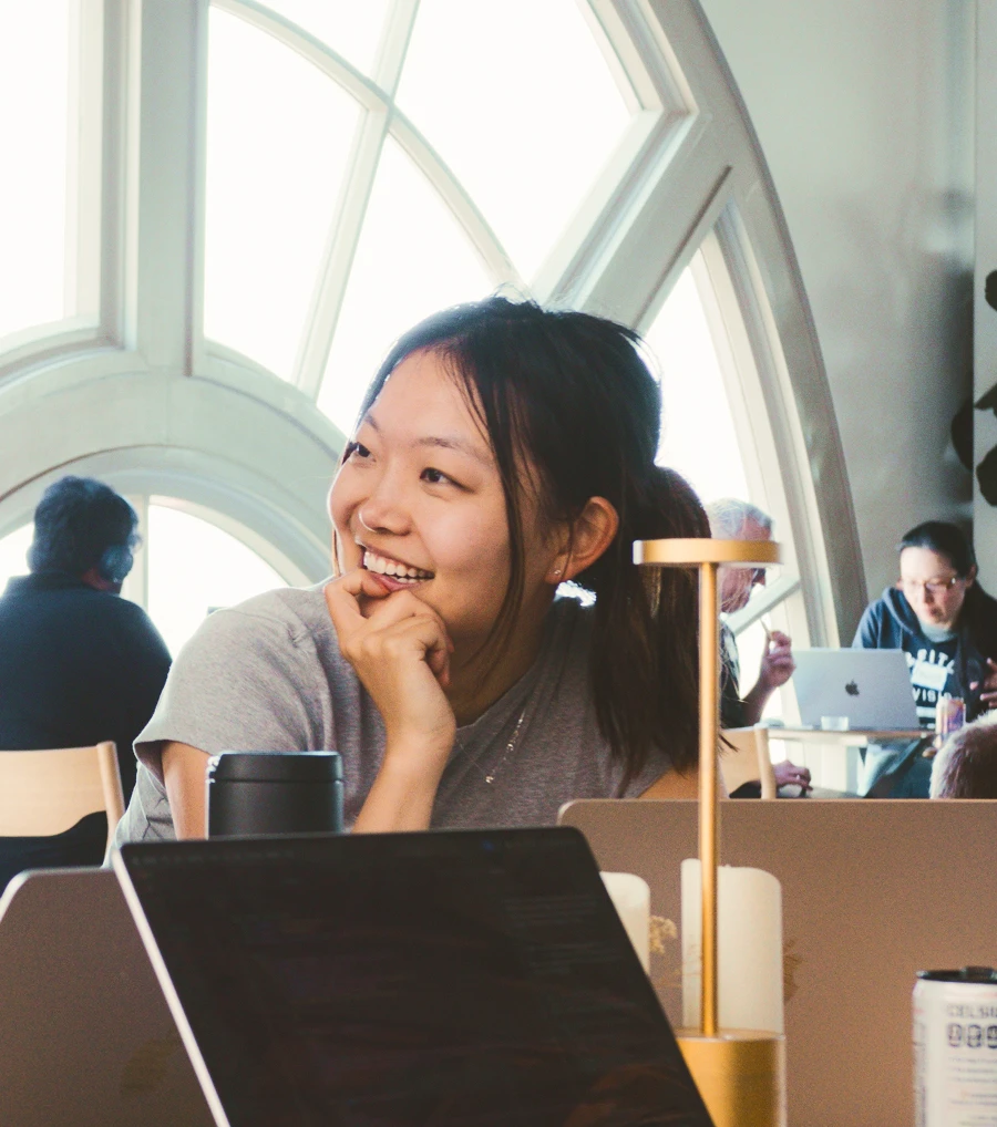 A person is smiling and looking to the side while seated at a table with laptops, a tumbler, and a small desk lamp in front of them. They are wearing a gray shirt and resting their chin on their hand. In the background, other people are also working on laptops in a bright room with a large arched window.