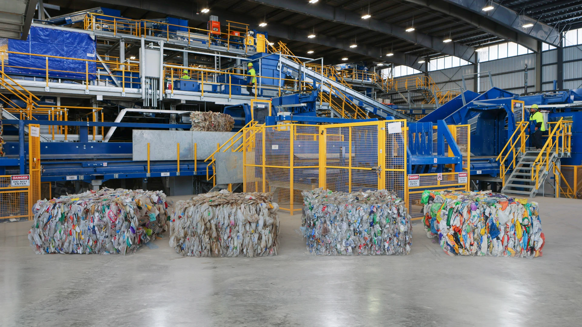 床に整然と並ぶ選別済みプラスチック廃棄物の圧縮梱包と大きな機械を備えた産業用リサイクル施設。作業者は高所のプラットフォームから作業を監督している