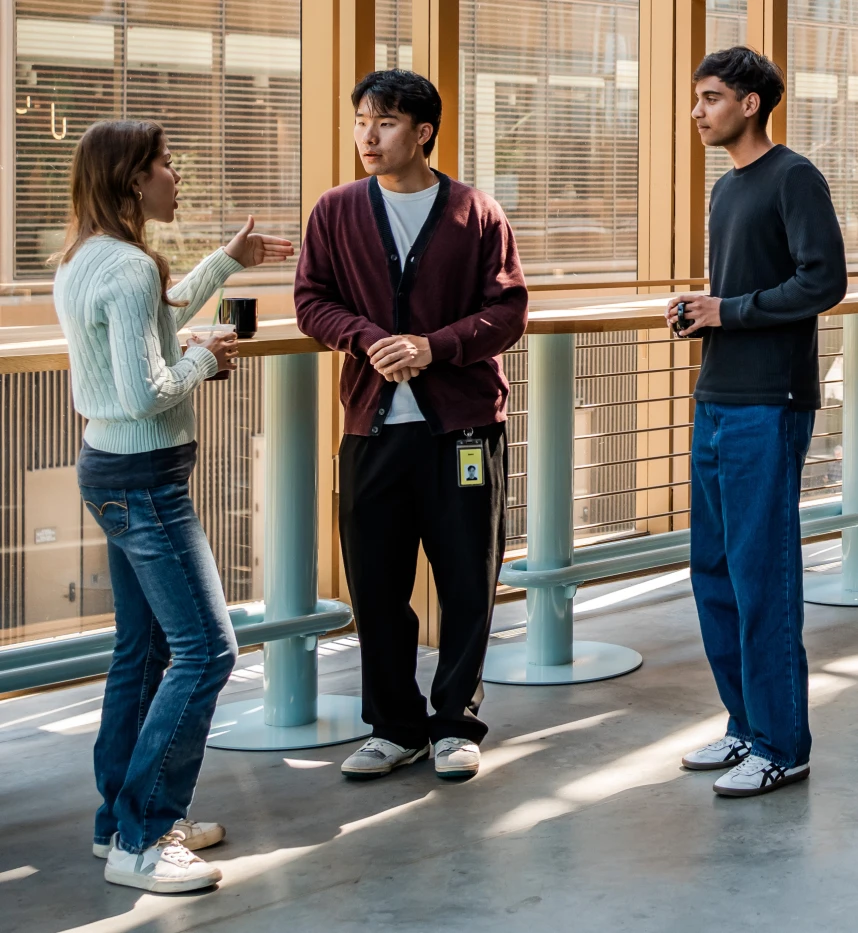 Three people standing and talking casually by a railing in a sunlit indoor space, two holding coffee cups.