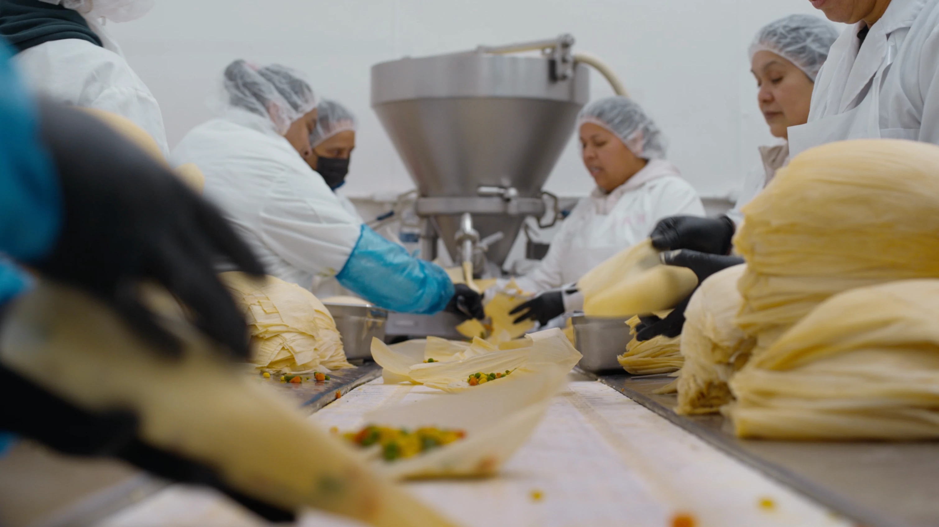 Workers wearing hairnets and gloves assemble tamales on a production line, stacking corn husks and filling them with ingredients in a food processing facility.