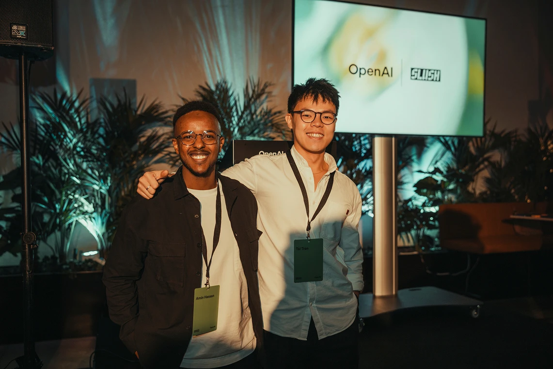 Two people smiling and posing together at an event. They both wear lanyards with name badges; the one on the left has a badge reading “Amin Hassan,” and the one on the right has a badge reading “Tai Tran.” Behind them is a large display screen showing the logos of “OpenAI” and “Slush,” with plants and event lighting in the background.