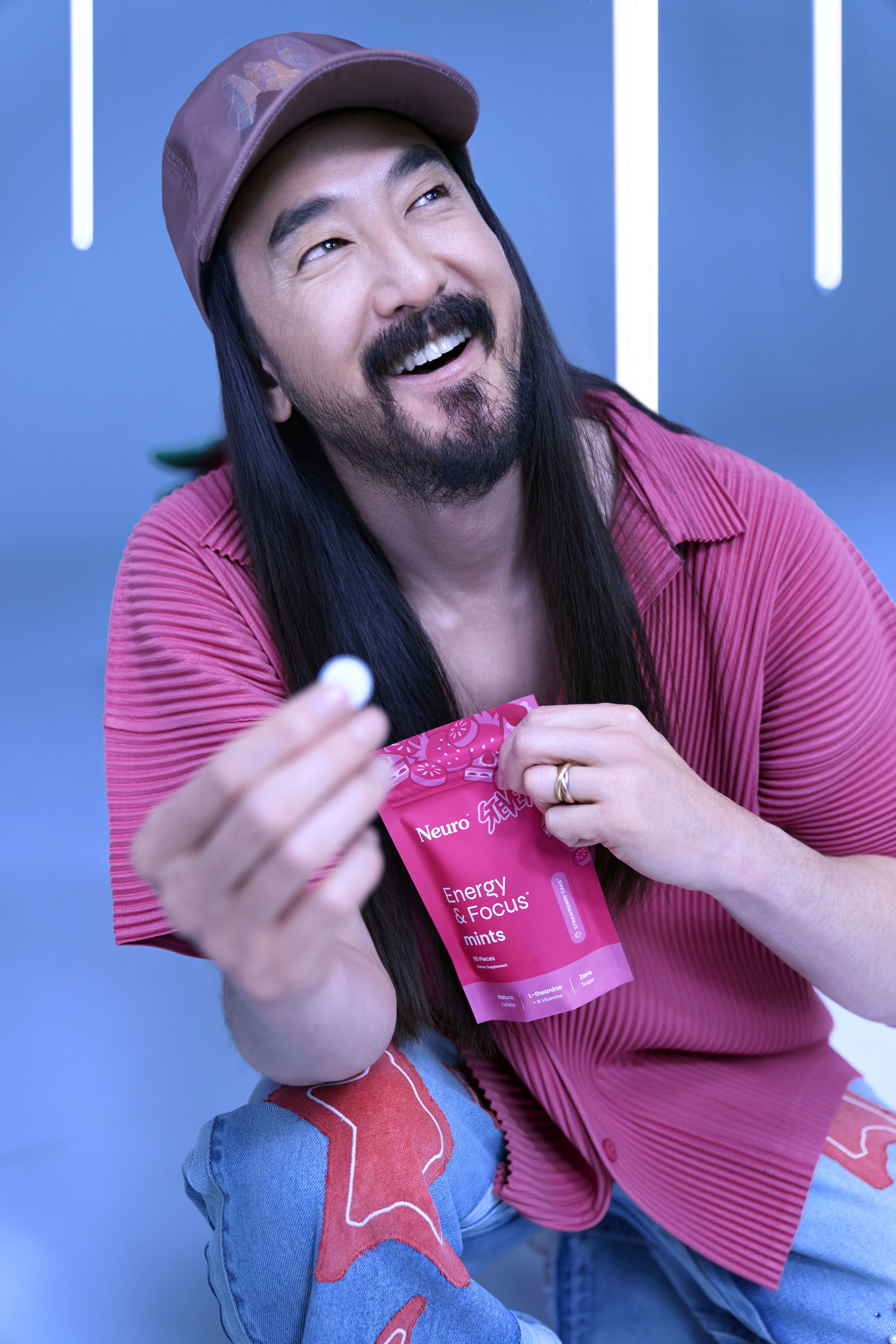Person smiling and holding a piece of Neuro Energy & Focus mint while seated, wearing a pink shirt and cap, with a matching pink product pouch in hand.