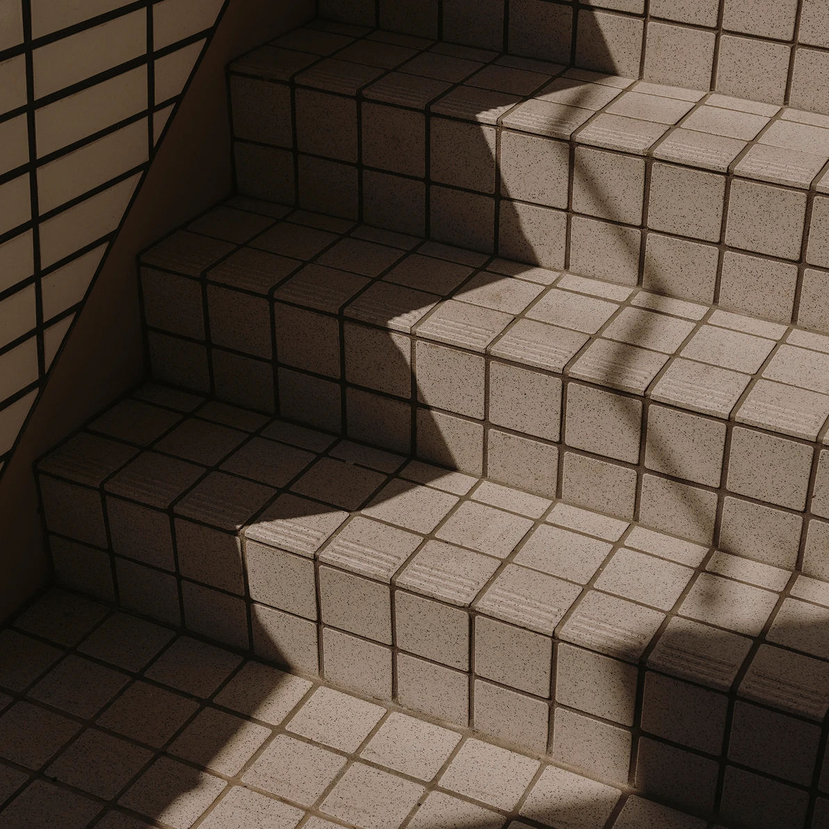A staircase made of square beige tiles, with strong geometric shadows cast by sunlight. The clean grid pattern and angled light create a minimal, architectural composition.