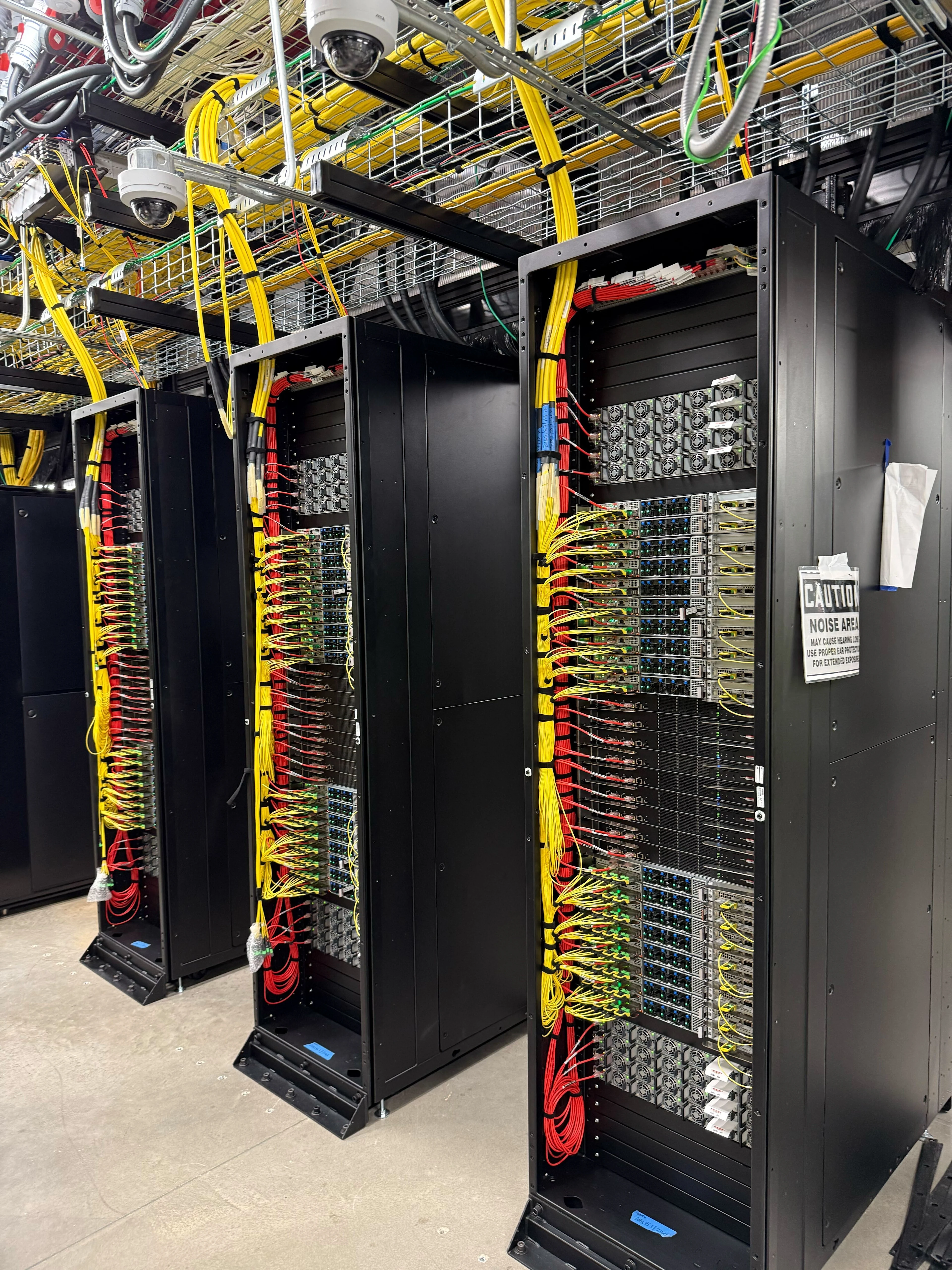 Filas de racks de servidores negros con cables de red amarillos y rojos en un centro de datos, con bandejas de cables aéreos y cámaras de seguridad visibles.