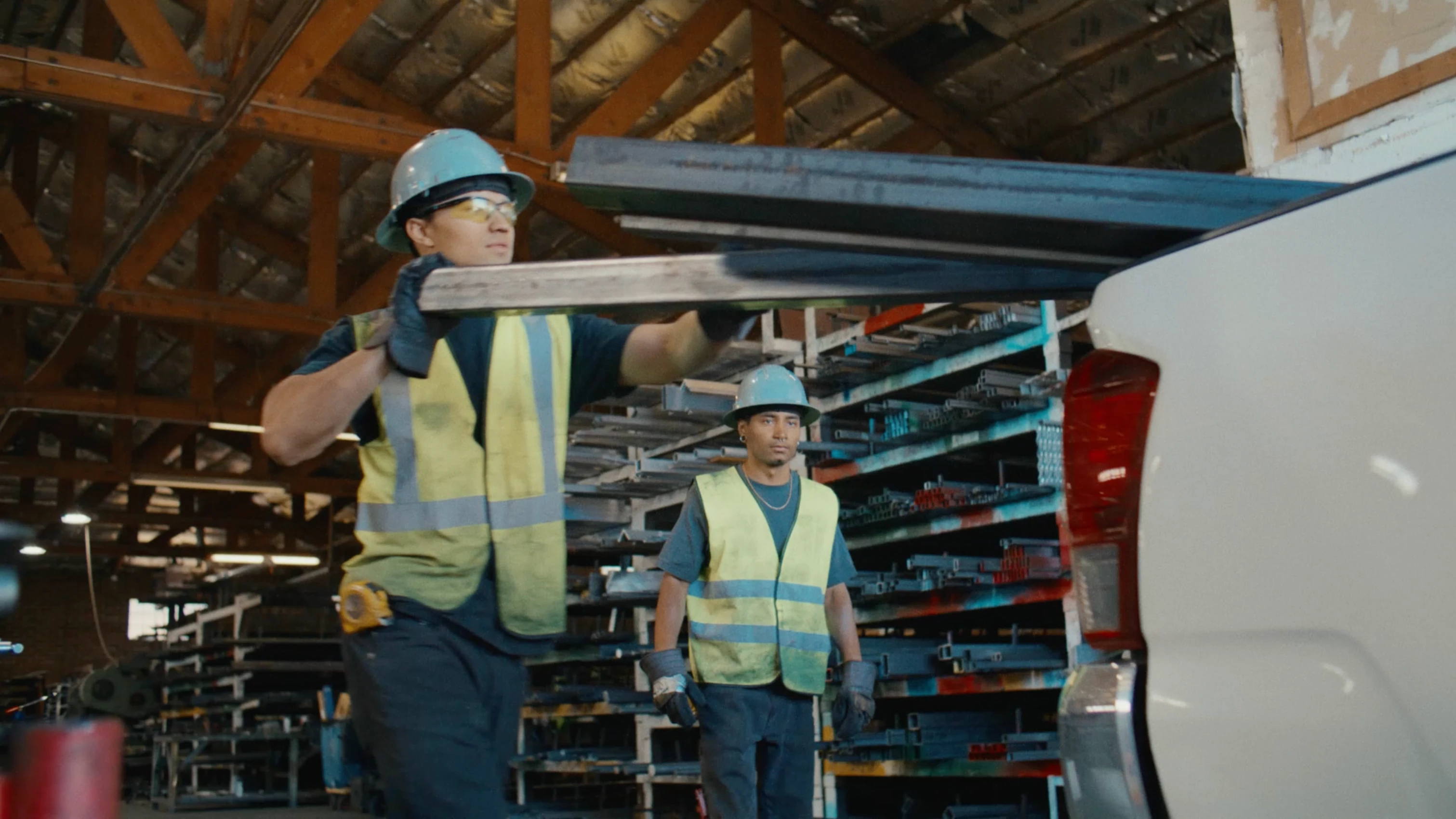 Two workers wearing hard hats and yellow safety vests carry long metal beams inside an industrial warehouse, loading them onto a truck.