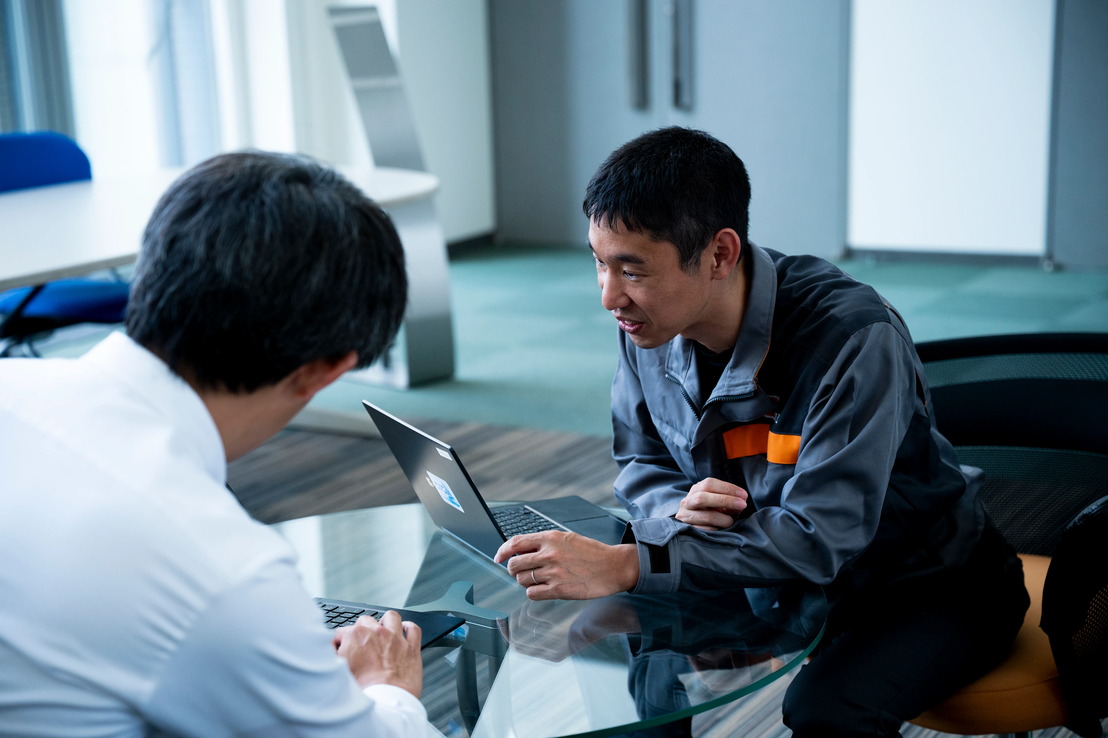 两名男子坐在玻璃桌旁，一边使用笔记本电脑一边讨论，其中一人穿着灰色工作服，另一人穿着白色衬衫。