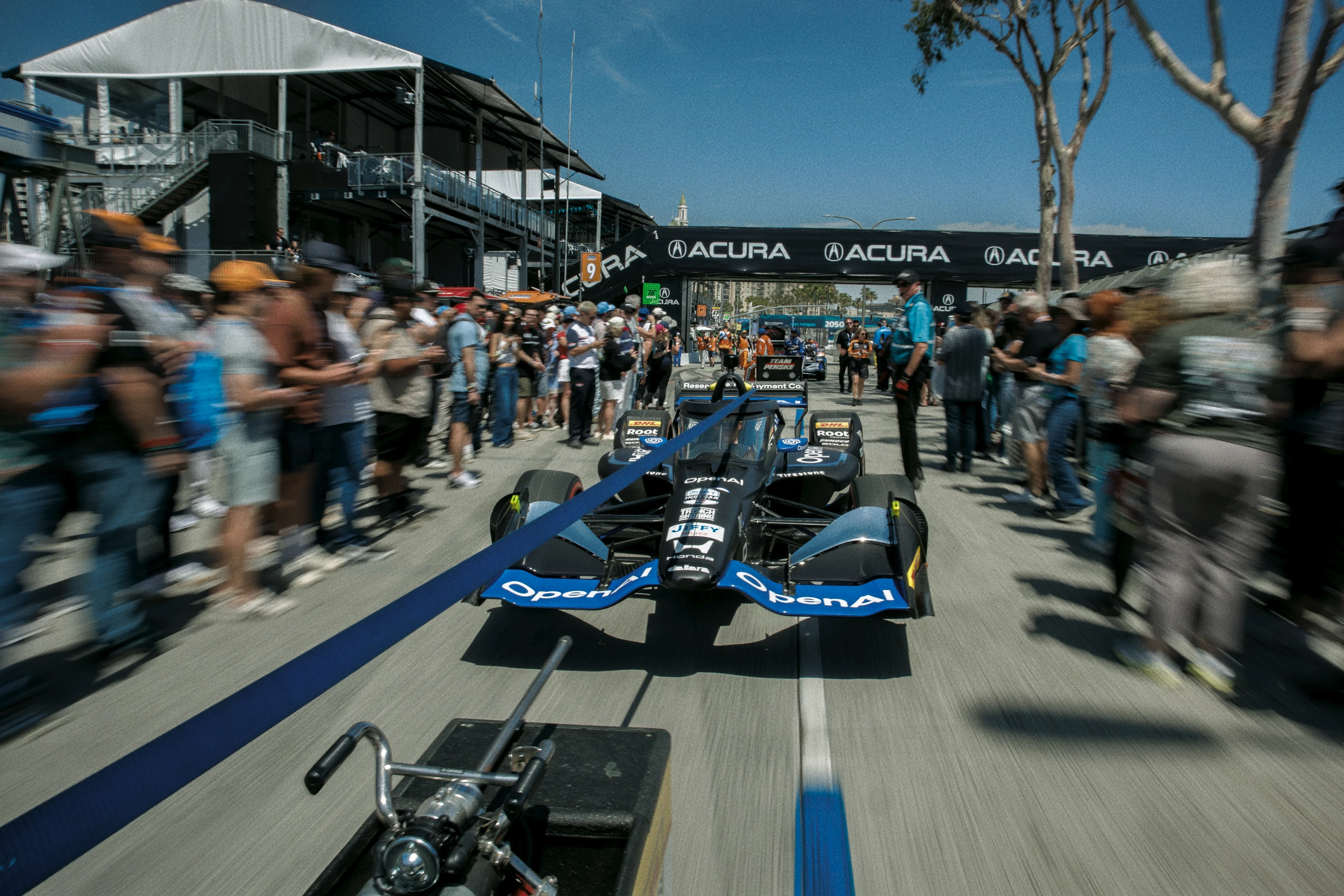 A blue IndyCar race car with OpenAI branding is being towed on a racing street circuit lined with spectators on both sides under clear blue skies. A black Acura banner spans across the track.