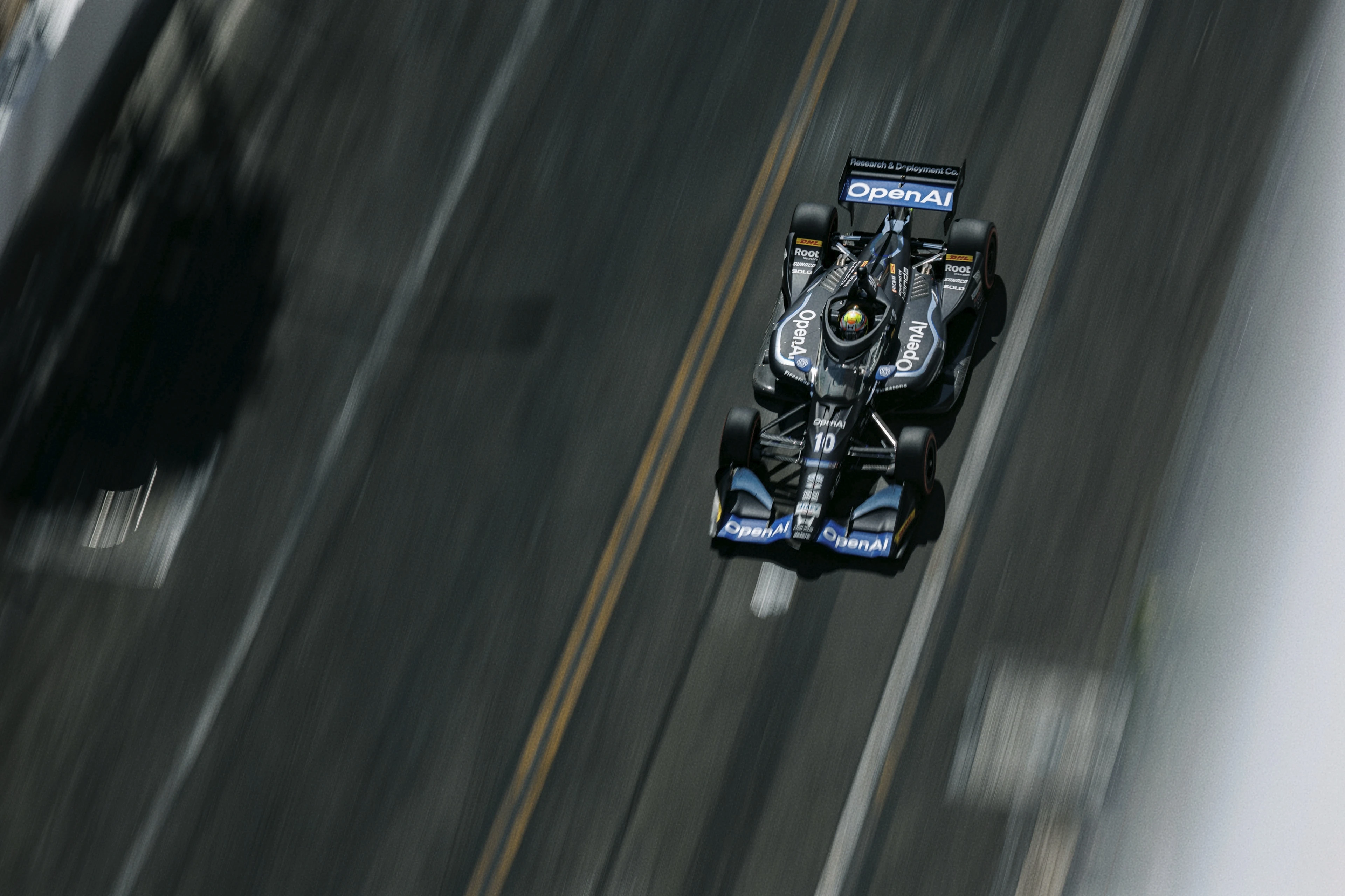 A black IndyCar race car with OpenAI branding speeds on a track, viewed from above, with motion blur emphasizing its fast movement.