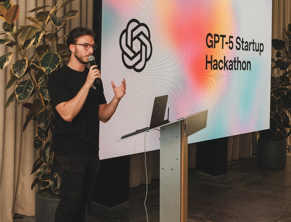 A person stands on stage speaking into a microphone beside a podium with a laptop. Behind them is a large screen displaying the OpenAI logo and the words “GPT-5 Startup Hackathon” on a colorful gradient background. Potted plants and curtains frame the space.