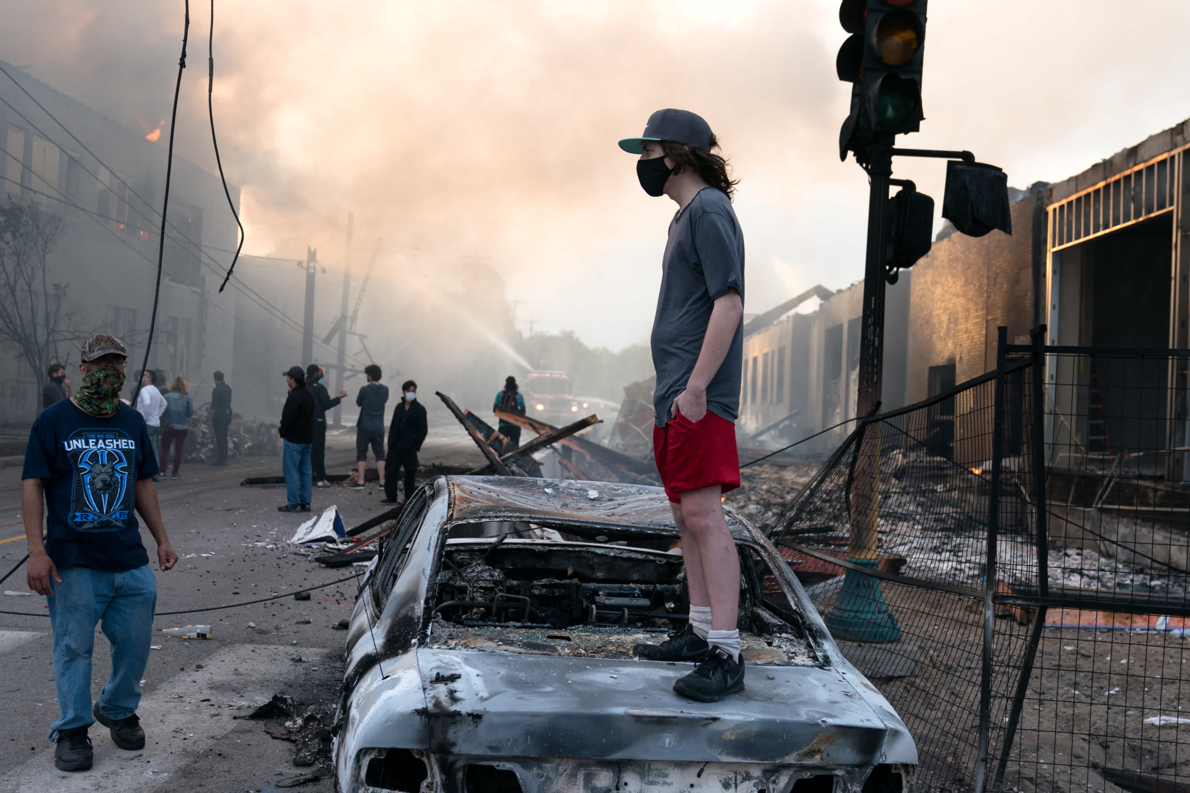 A man stands on a burned out car on Thursday morning as fires burn behind him in the Lake St area of Minneapolis, Minnesota
