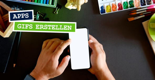 Hände halten ein Smartphone über einen Schreibtisch, der mit Farben, Stiften und Pinseln bedeckt ist.