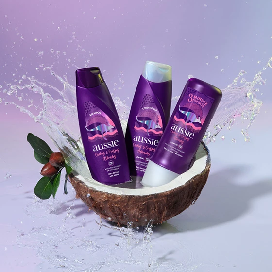 Frascos da linha Aussie Cachos & Crespos atividades dentro de meio coco aberto com fundo lilás.