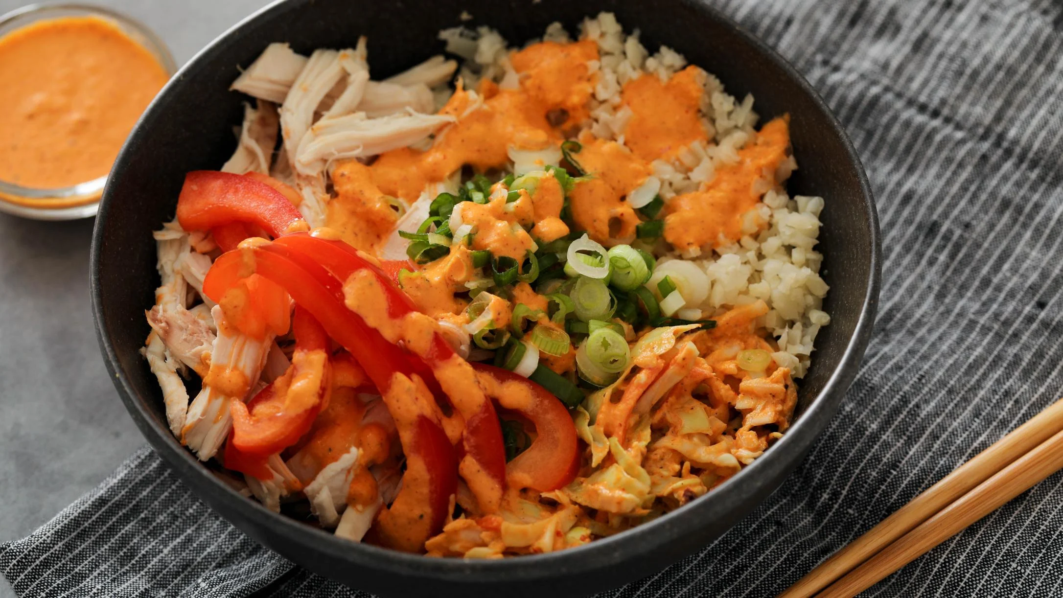 Black bowl with rice, shredded chicken, red bell peppers, green onions, and orange sauce, chopsticks nearby.
