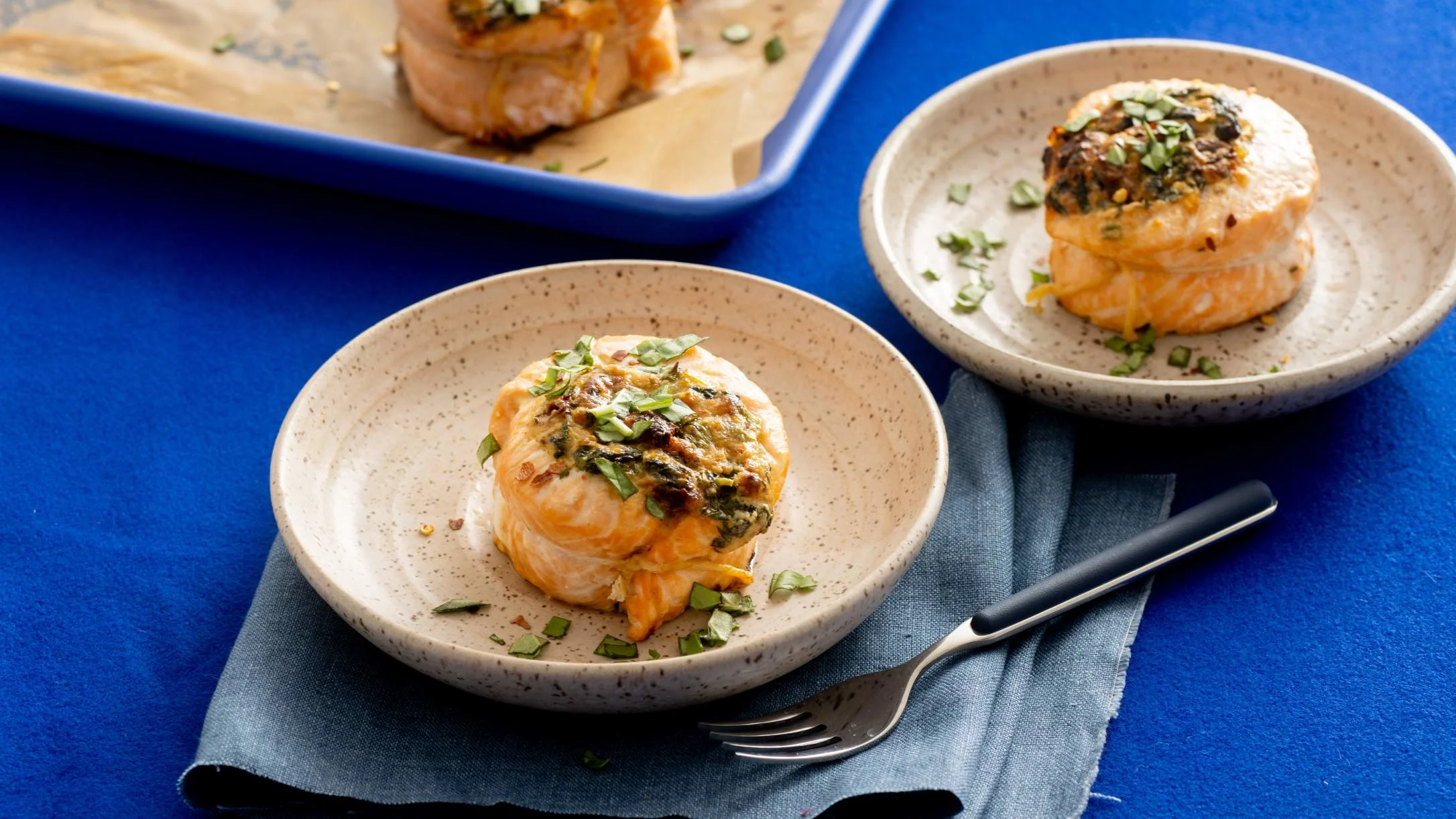 Herb-topped salmon medallions served on speckled ceramic plates against a vibrant blue background with a silver fork.