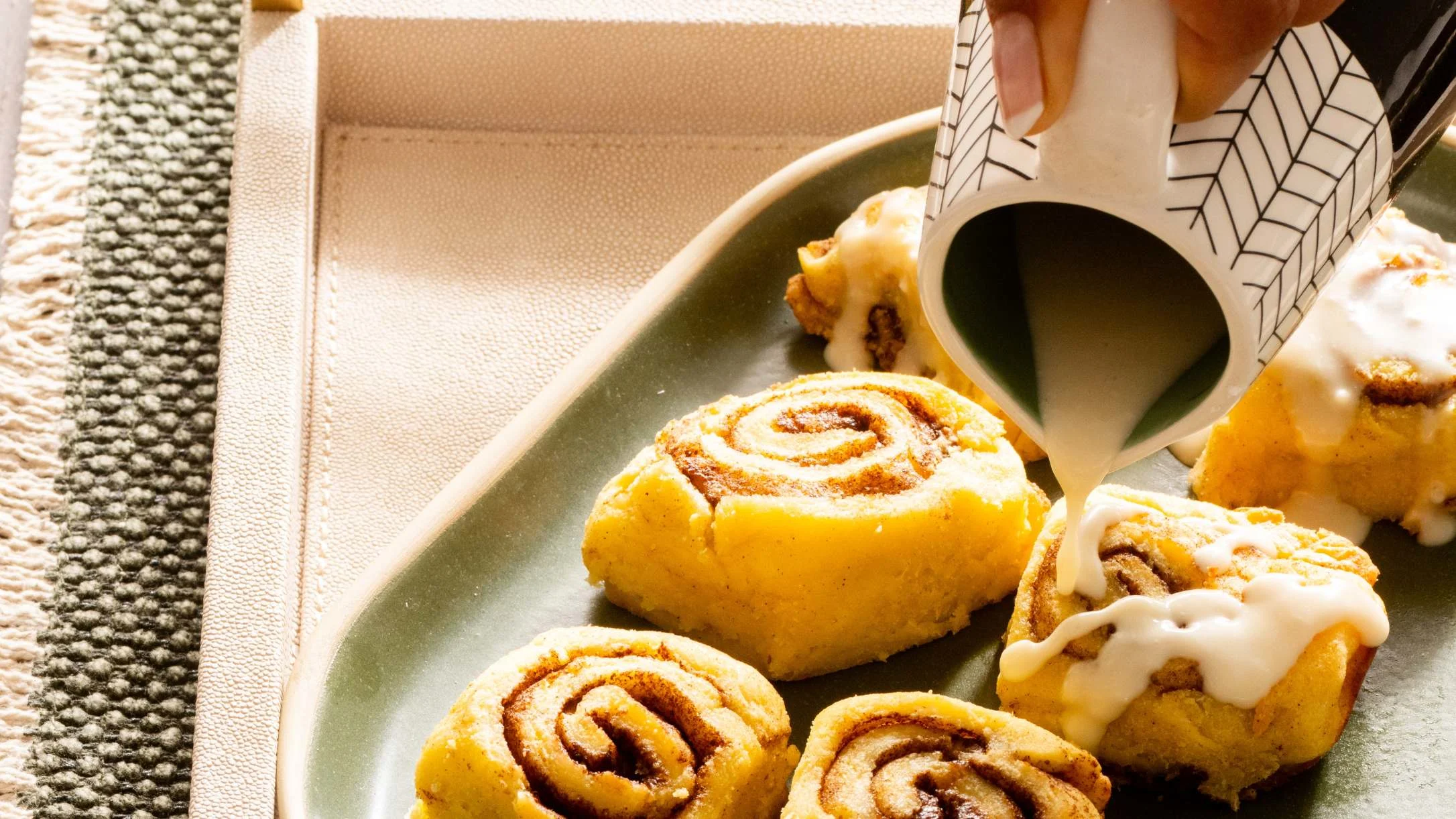 A hand pours white icing from a small pitcher over golden cinnamon rolls on a green serving platter.