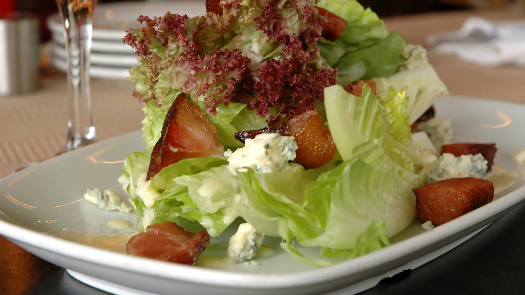 A fresh salad with green and red lettuce, bacon, goat cheese crumbles, and dressing on a white plate.