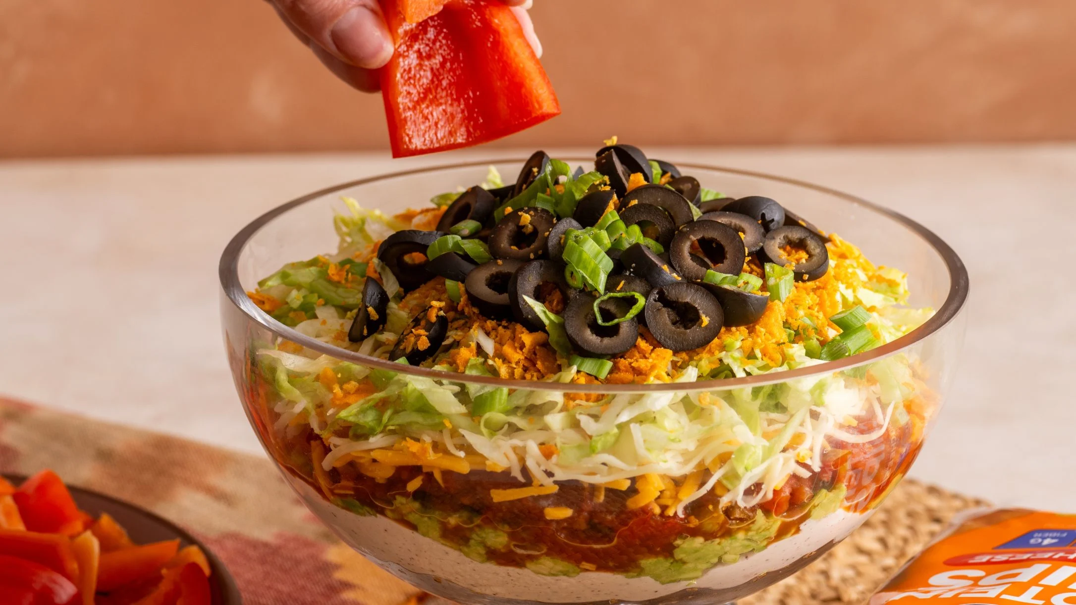 Hand dipping red bell pepper into a layered taco dip bowl topped with black olives, green onions, and shredded cheese.