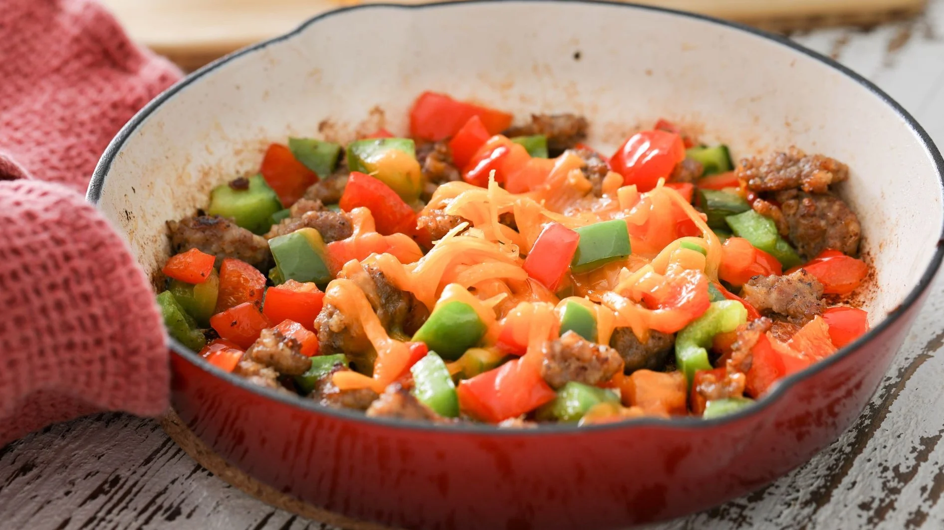 Skillet meal with sausage, diced red peppers, green bell peppers, and melted cheese in a red cooking pan.