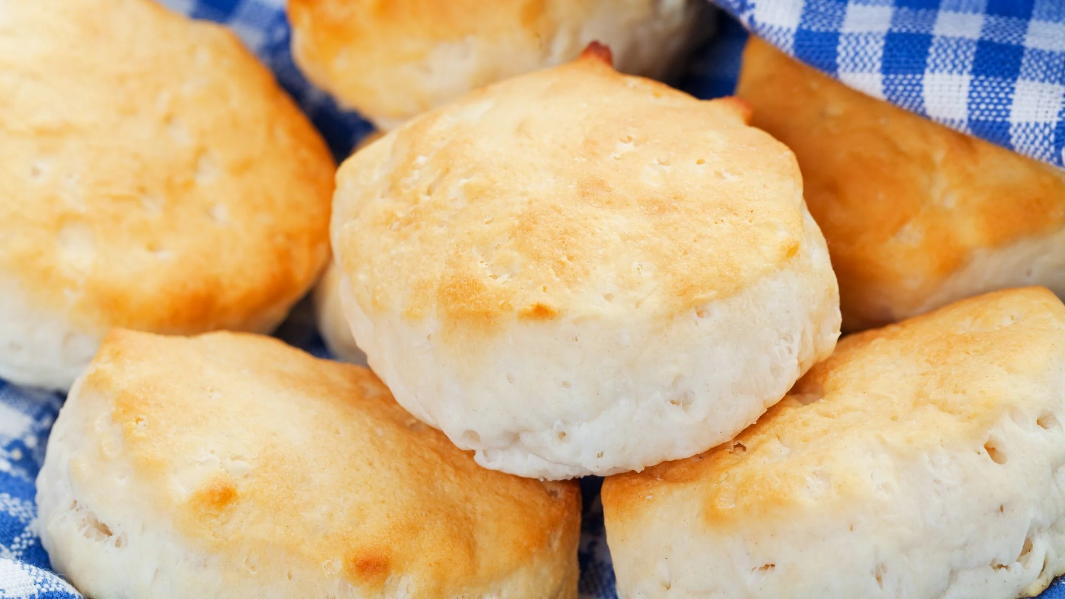 Freshly baked biscuits with golden tops stacked on a blue and white checkered cloth.
