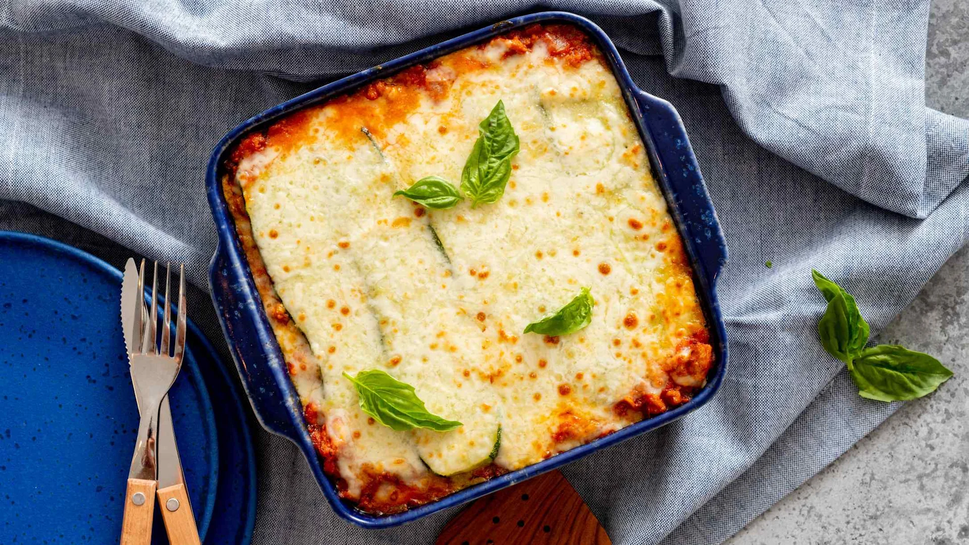 Cheesy lasagna in a blue baking dish, topped with golden melted cheese and fresh basil leaves, beside a fork and knife.