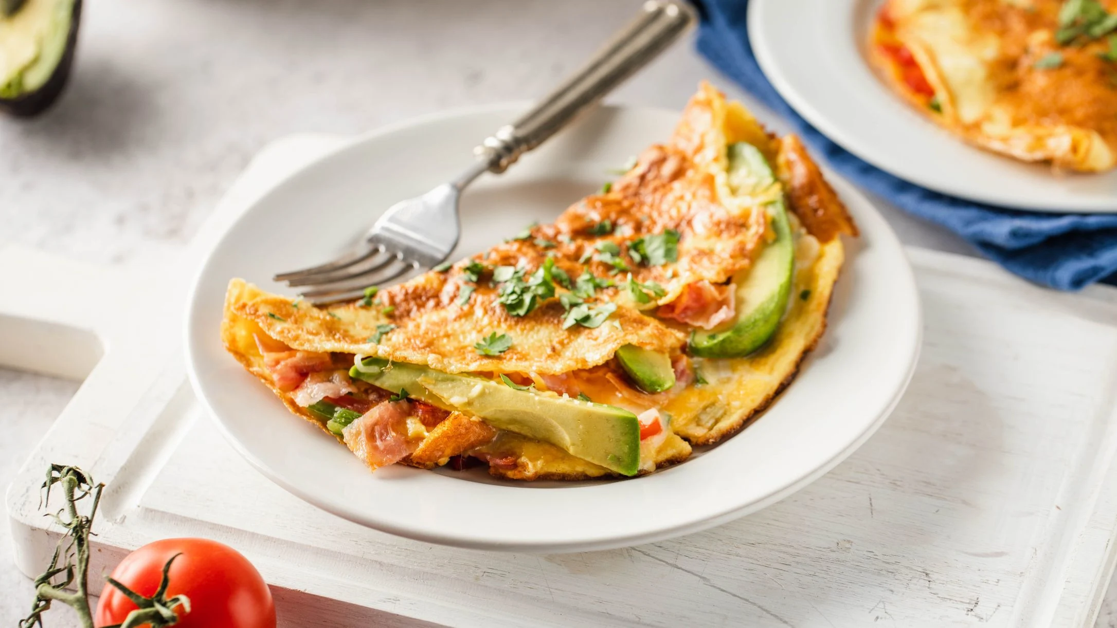 Folded omelet with avocado slices and herbs on white plate, fork beside it, tomato in corner of white table.