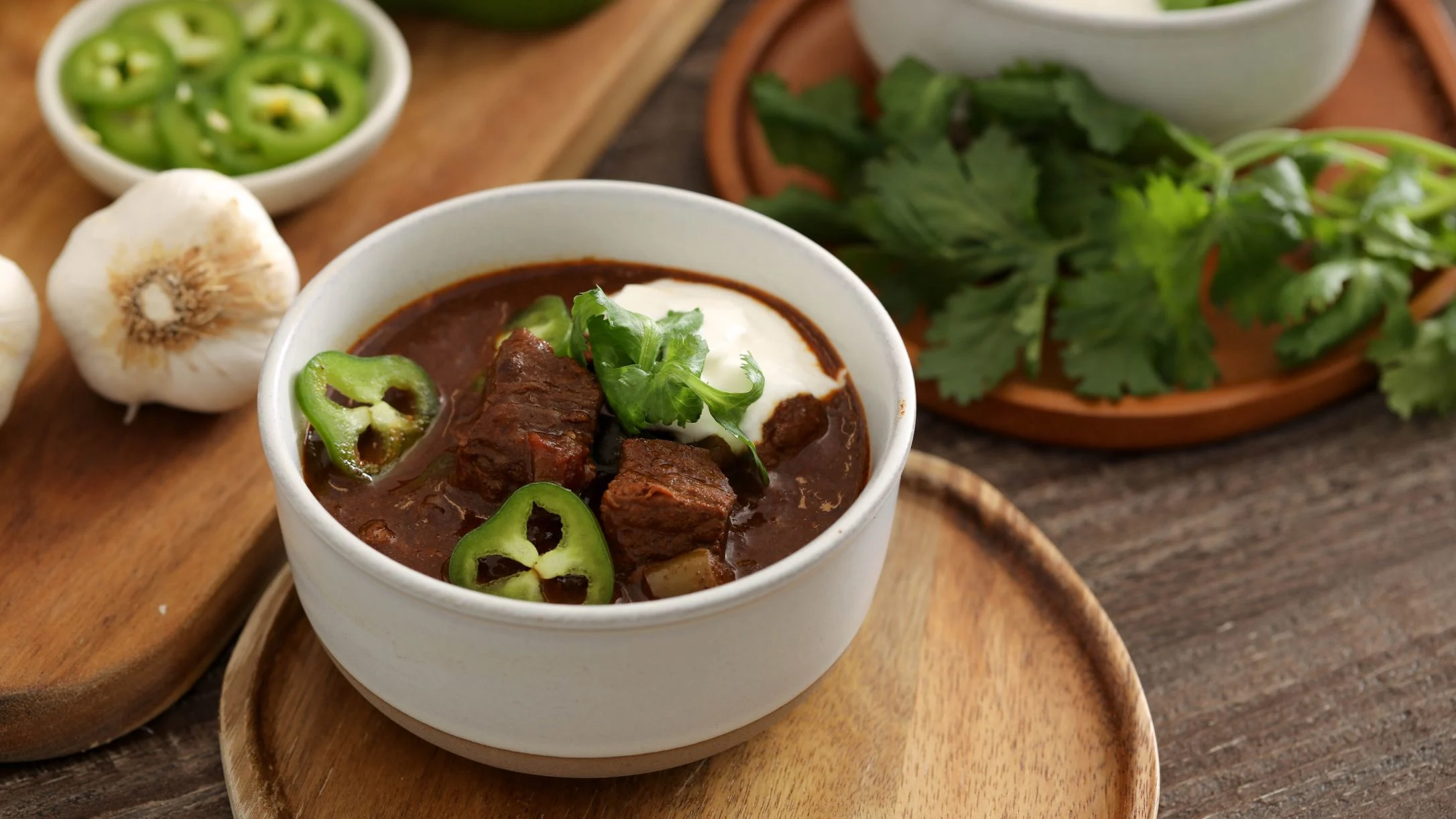 A white bowl of beef chili topped with jalapeño slices, sour cream, and cilantro, surrounded by garlic, fresh herbs, and peppers.