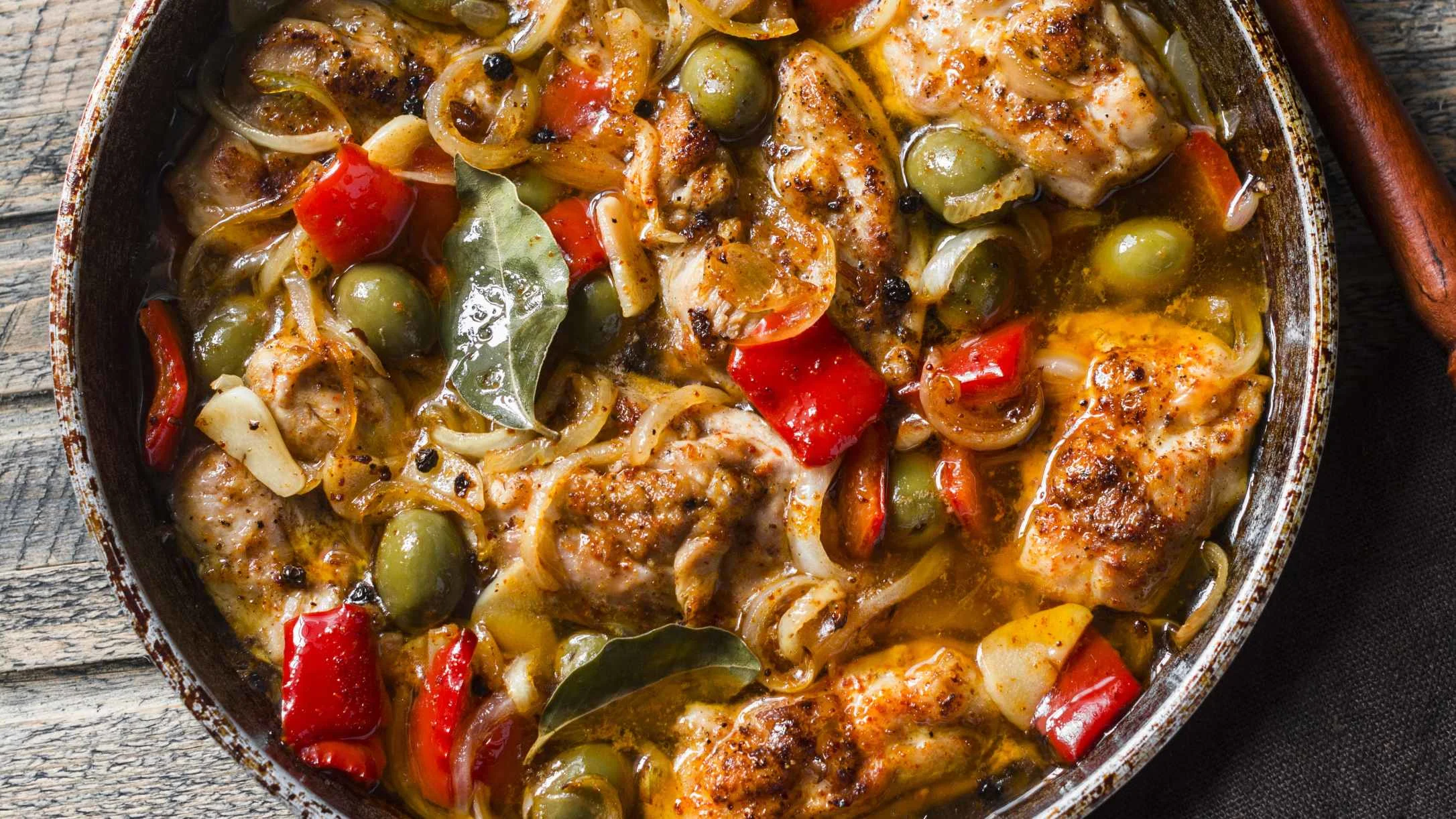 Braised chicken with green olives, red peppers, onions, and bay leaves in a rustic pan with golden sauce.