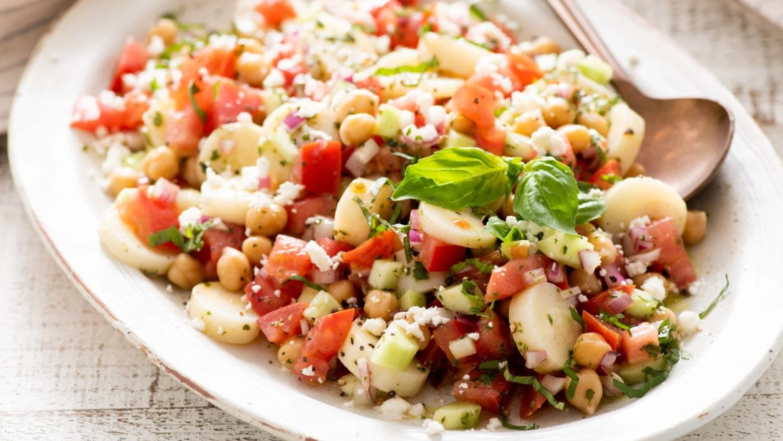 Fresh Mediterranean bean salad with tomatoes, chickpeas, herbs, and feta cheese garnished with basil leaves on a white plate.