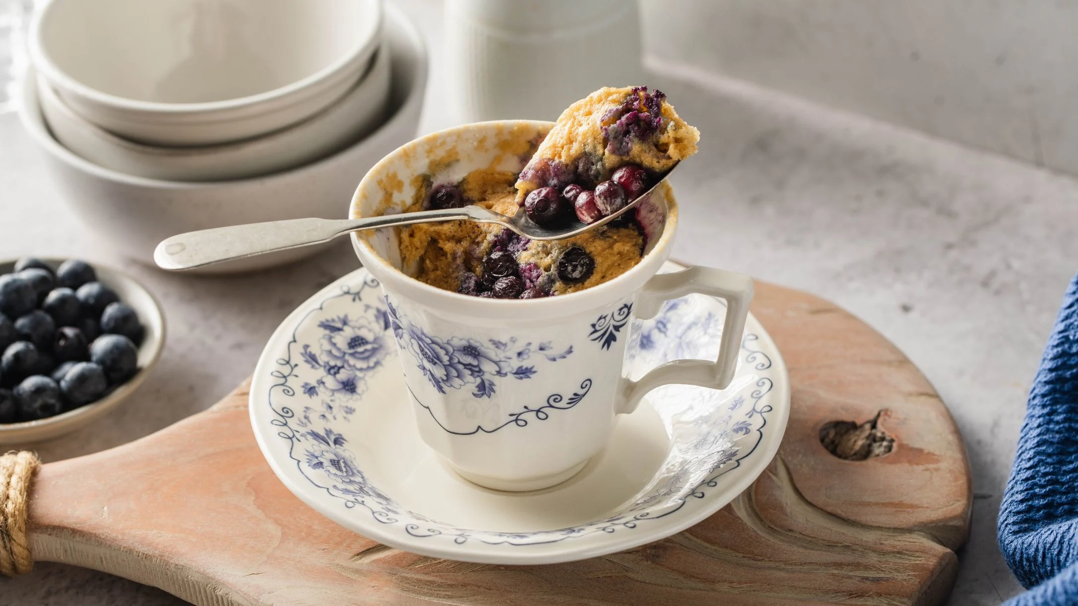 Blueberry mug cake in decorative porcelain teacup with spoon, fresh blueberries nearby on wooden board.