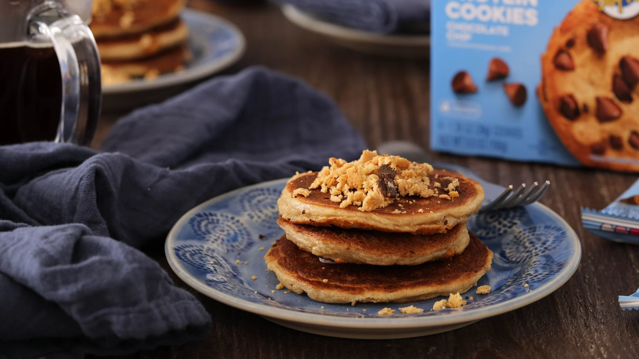 Stack of fluffy pancakes topped with crumbled chocolate chip protein cookies on a blue plate, with syrup and cookie box nearby.