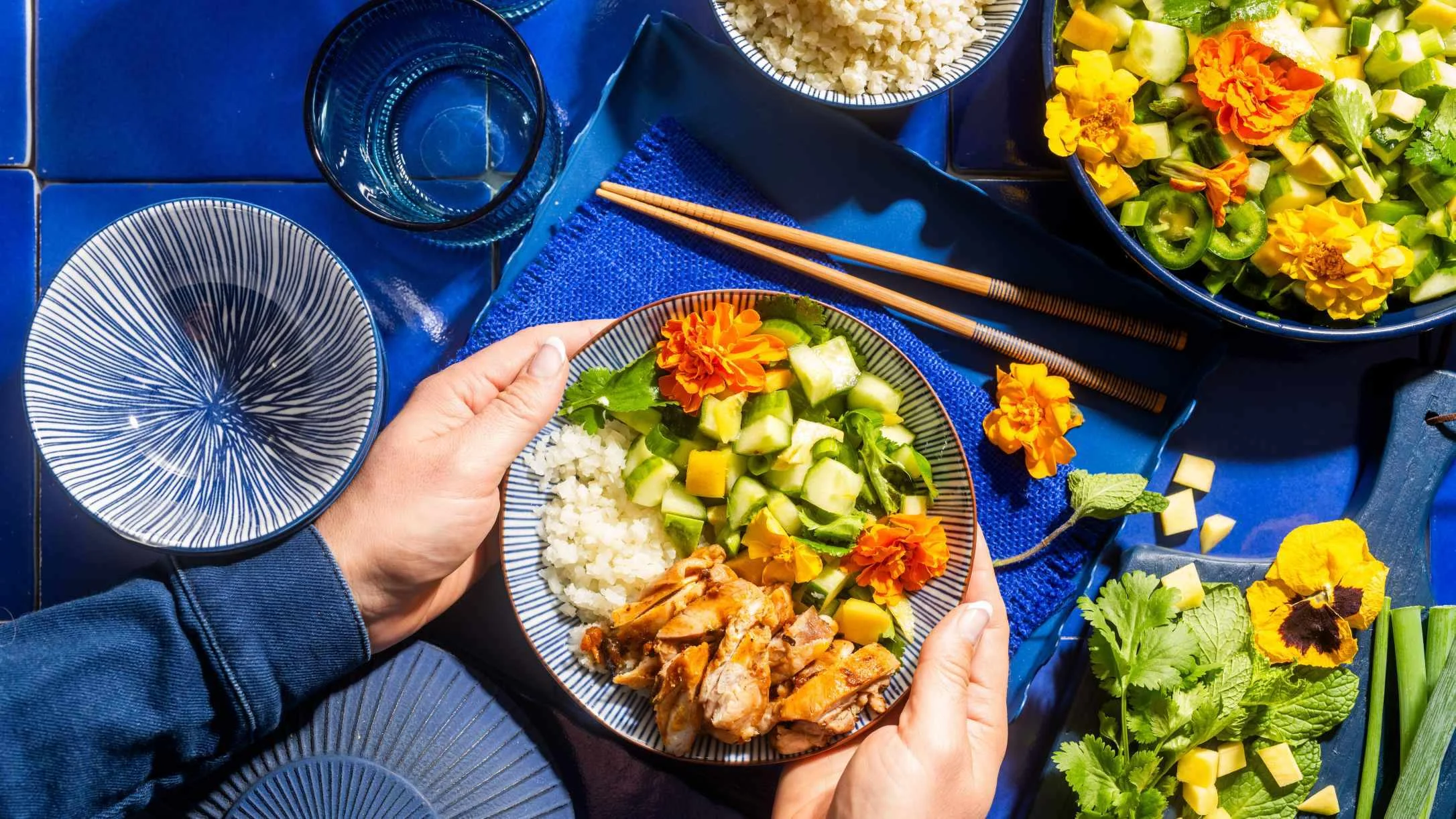 Hands holding a bowl with cauliflower rice, chicken, and vegetables garnished with edible flowers on a vibrant blue table setting.