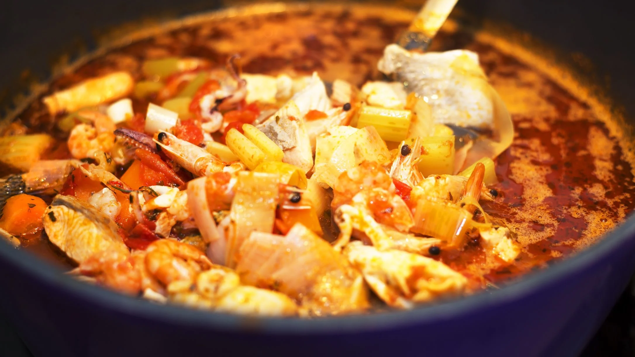 A rich seafood stew with shrimp, squid, fish, and vegetables simmering in a red tomato broth inside a blue pot.