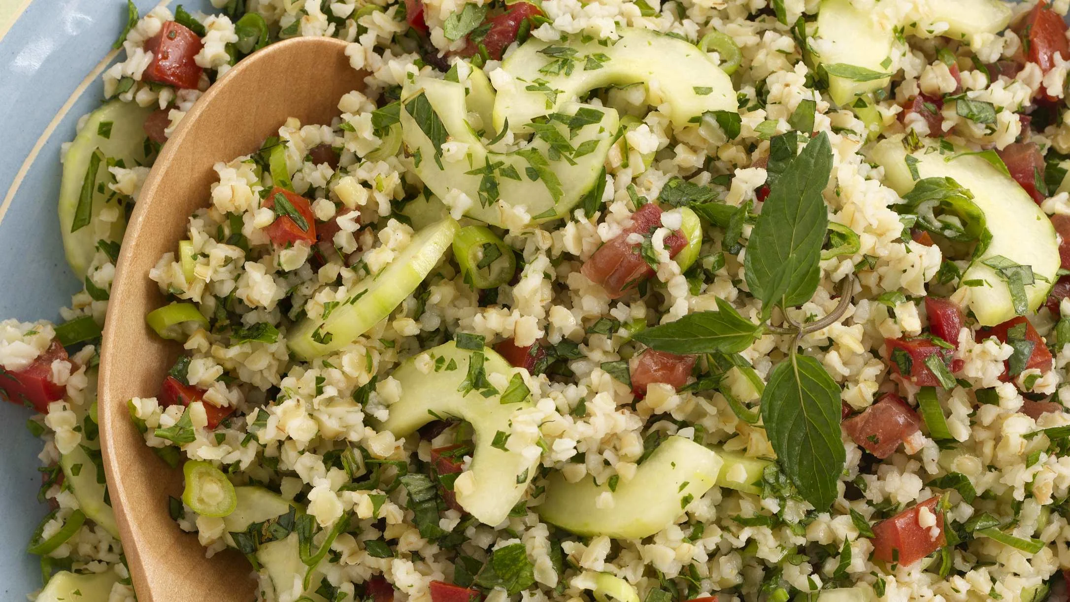 A close-up of tabbouleh salad with bulgur wheat, diced tomatoes, cucumber ribbons, and fresh herbs in a tan bowl.