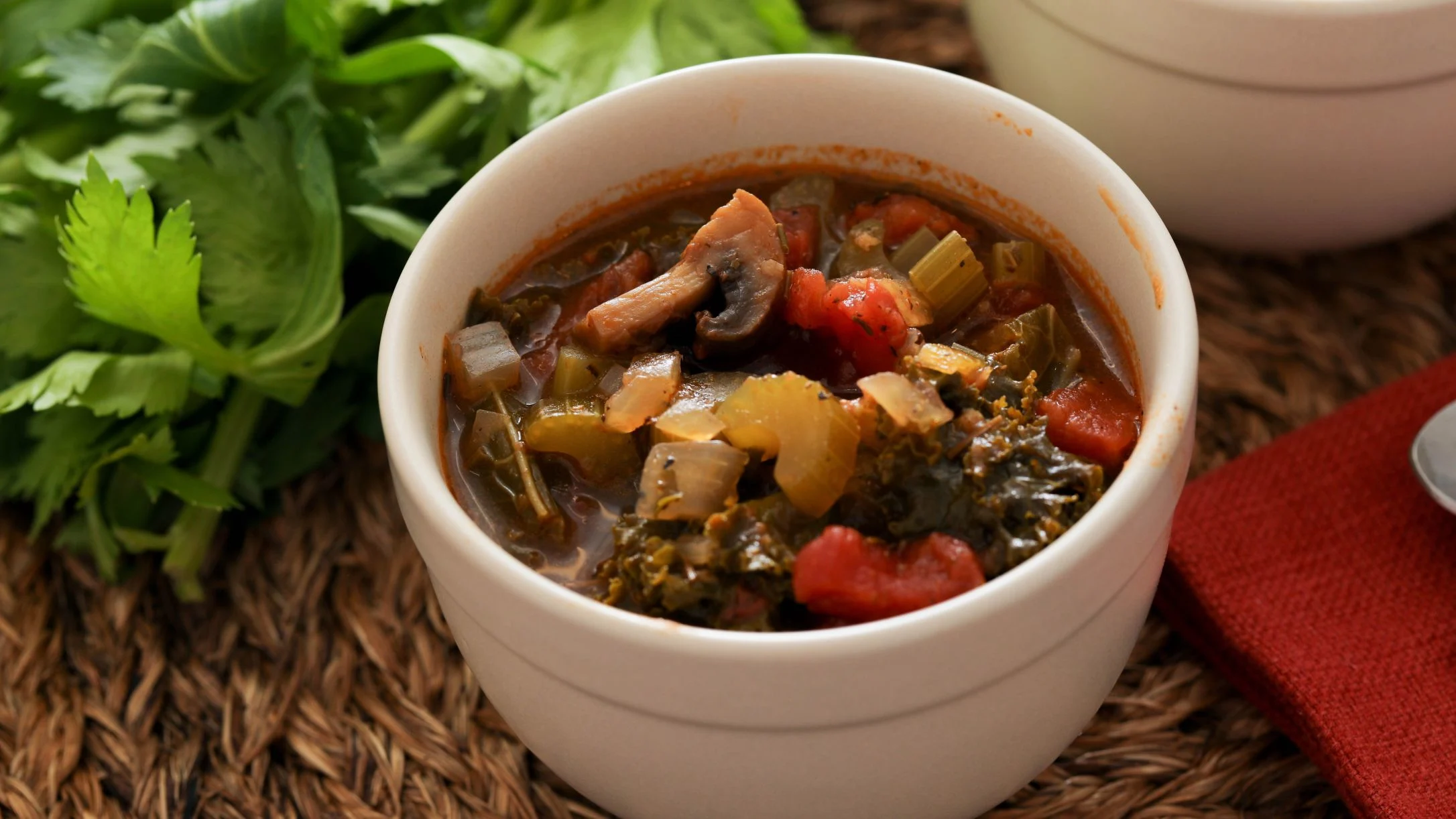 A white bowl of hearty vegetable soup with kale, tomatoes, mushrooms, and celery on a woven mat beside fresh celery.