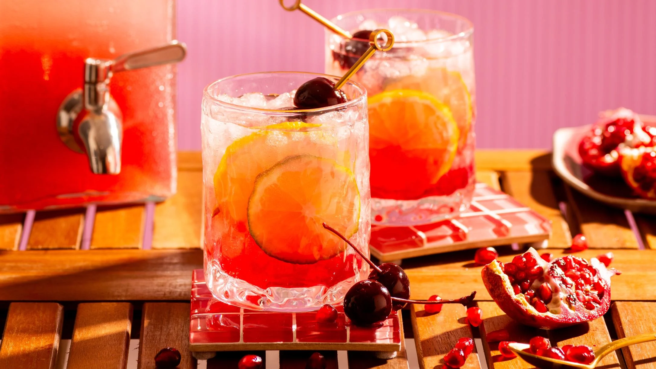 Two red cocktails with lemon slices, cherries, and ice on a wooden table, beside a drink dispenser and fresh pomegranate.